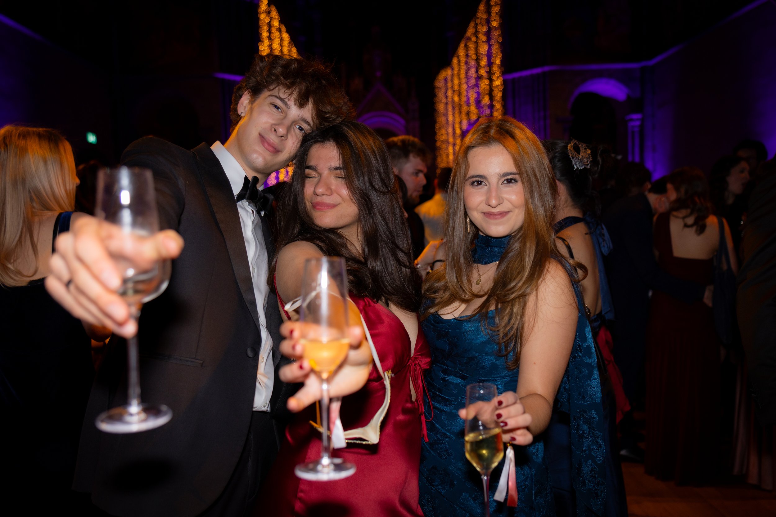 Group of young adults dressed in formal attire at a lively celebration or party, with a dimly lit background decorated with hanging string lights.