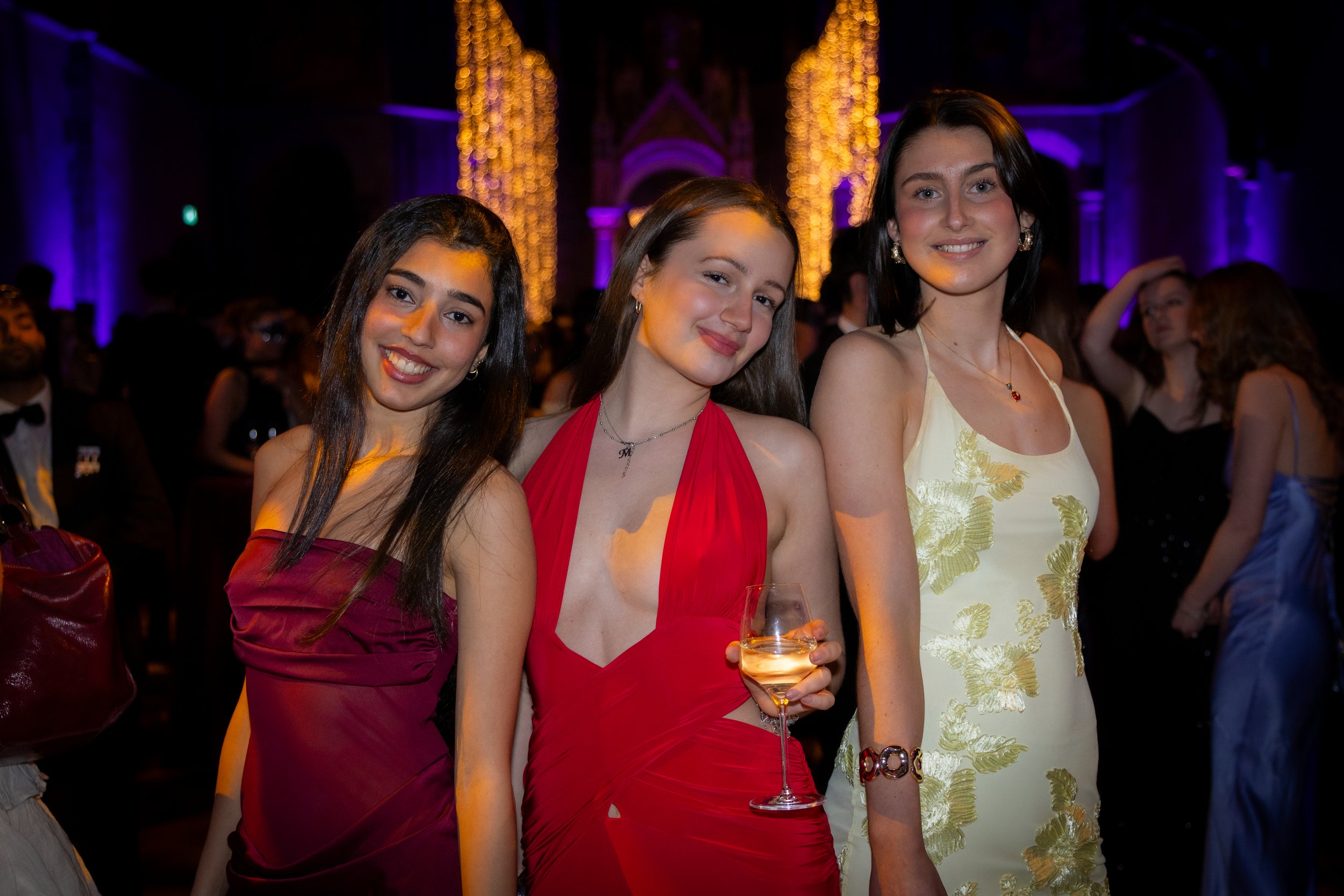 Three young women in elegant dresses posing at a formal event with bright lights and a grand venue in the background.