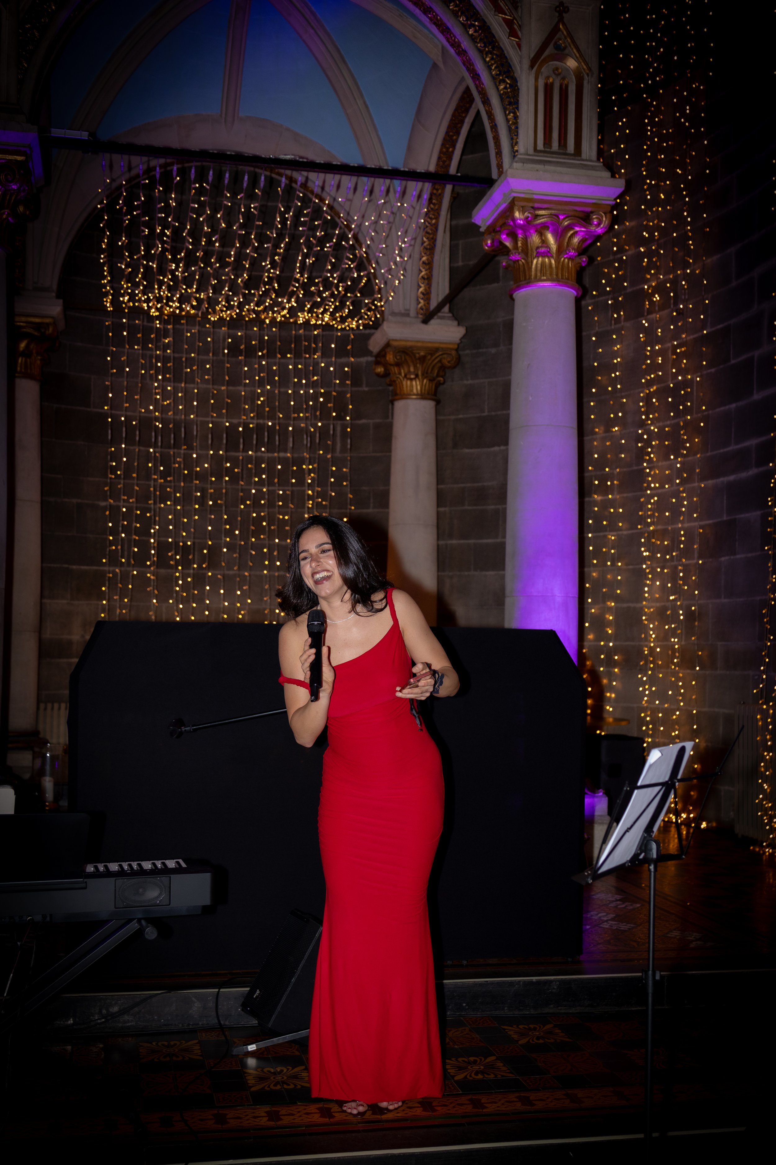 A woman in a red dress holding a microphone and standing infront of a piano with a band. The backdrop is a decorated venue with chandeliers and string lights.