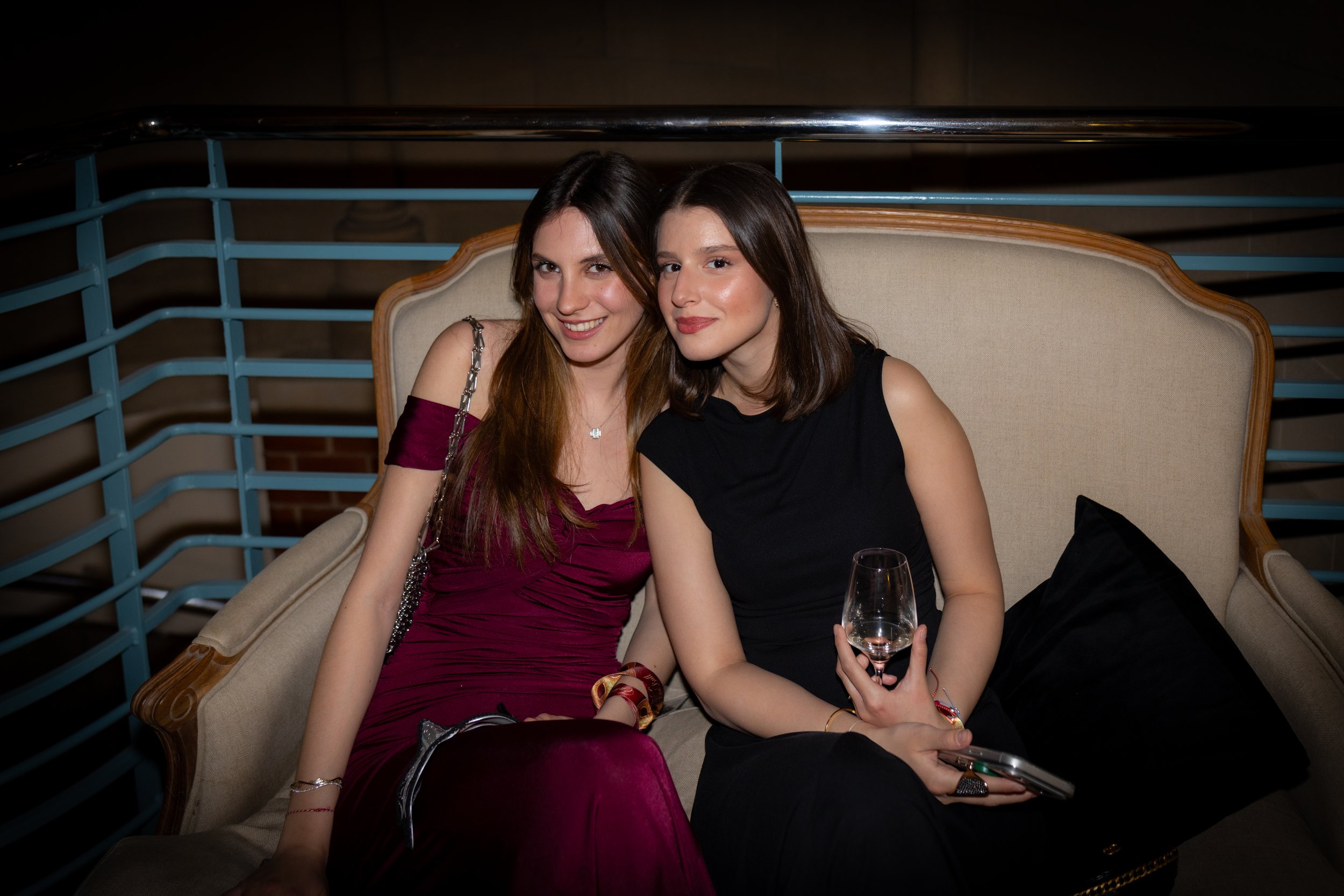 Two women sitting on a beige couch, smiling at the camera, one holding a wine glass, in a dimly lit room.