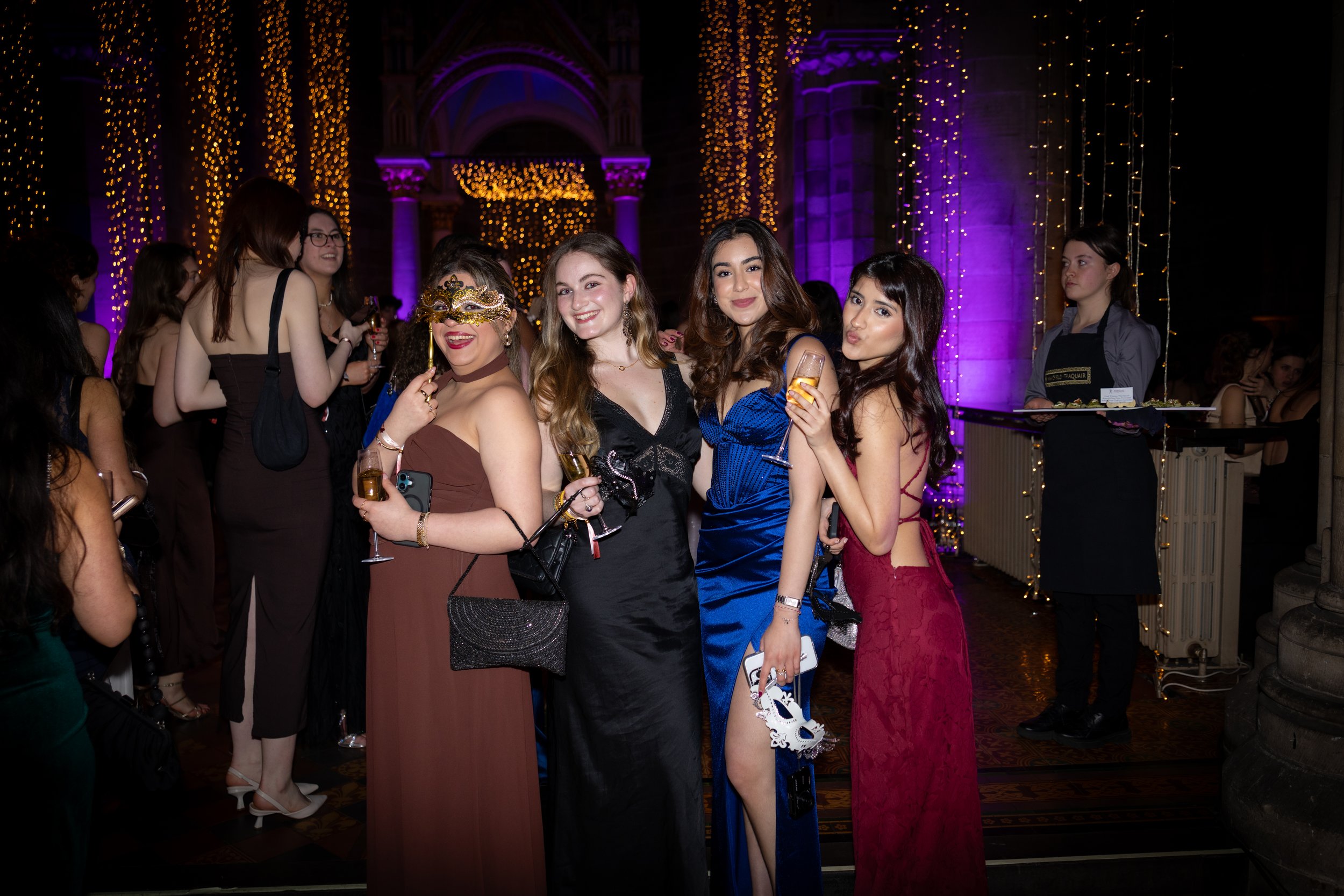 Group of women at a festive event, dressed in elegant gowns, with some wearing masks and holding drinks, surrounded by purple and yellow string lights.