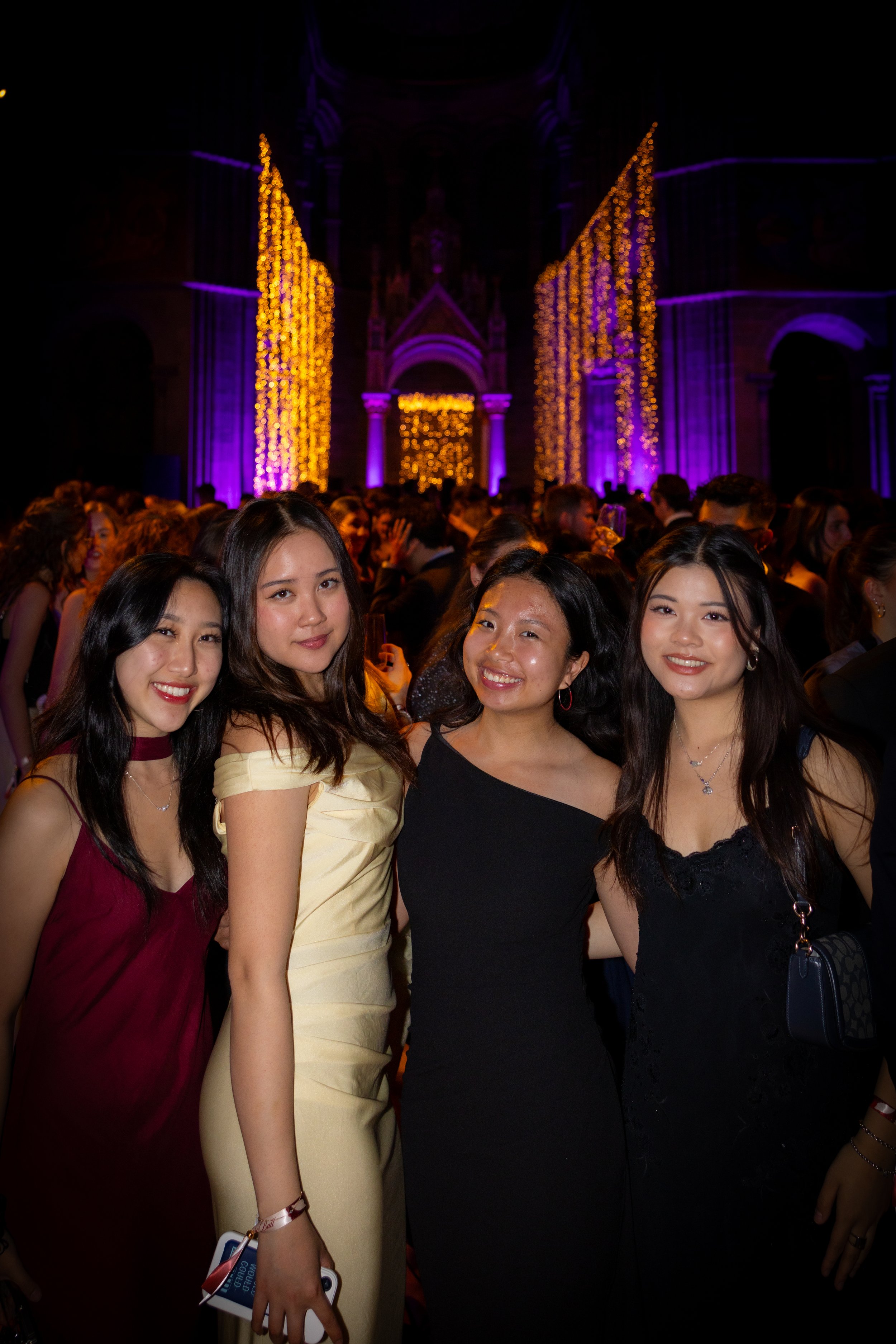 Four young women dressed in formal attire at a crowded event with purple and gold lights and decorations.