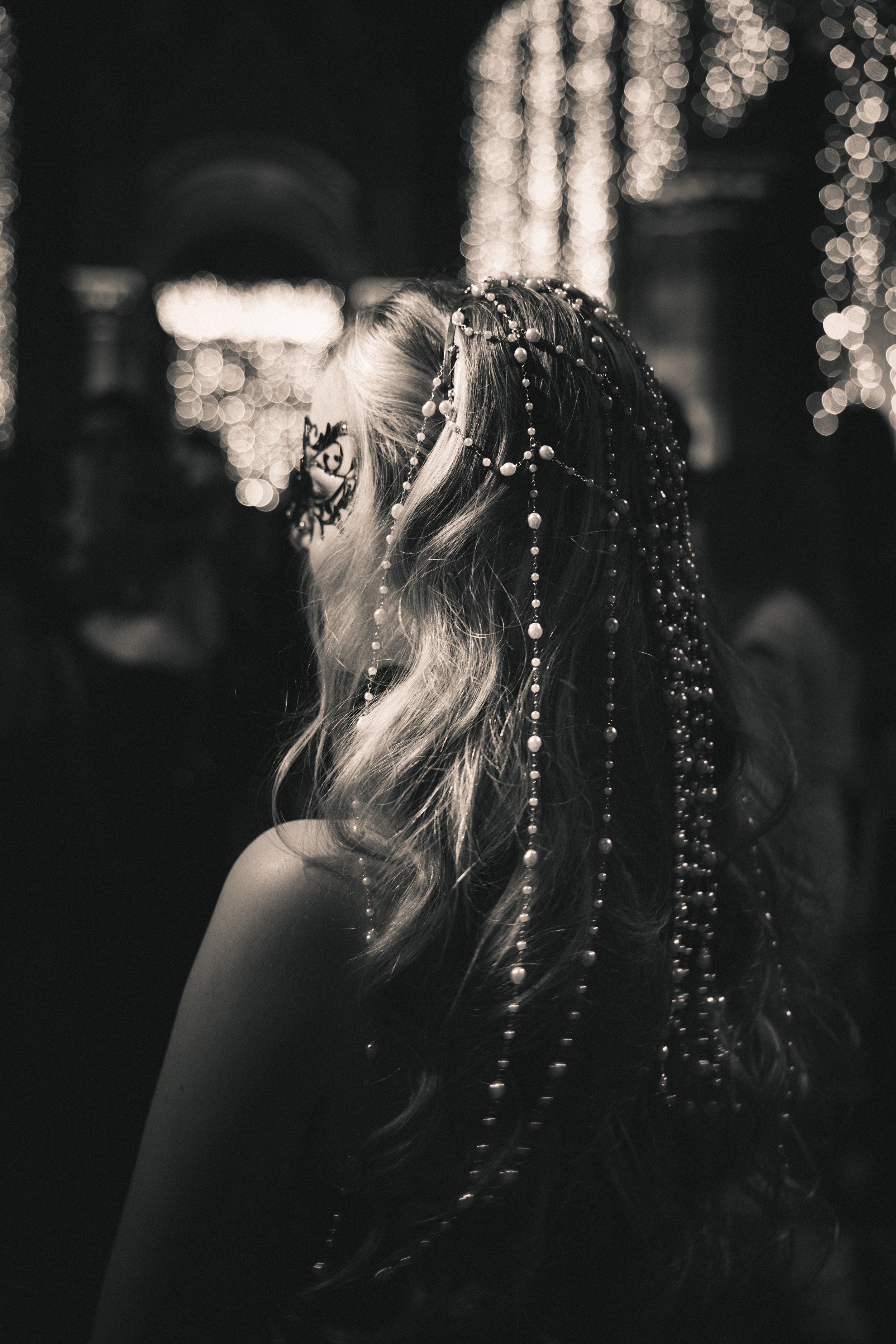 A woman with long, wavy hair decorated with pearls and beads, wearing glasses with floral designs, in front of a backdrop of out-of-focus string lights, in a black and white photo.