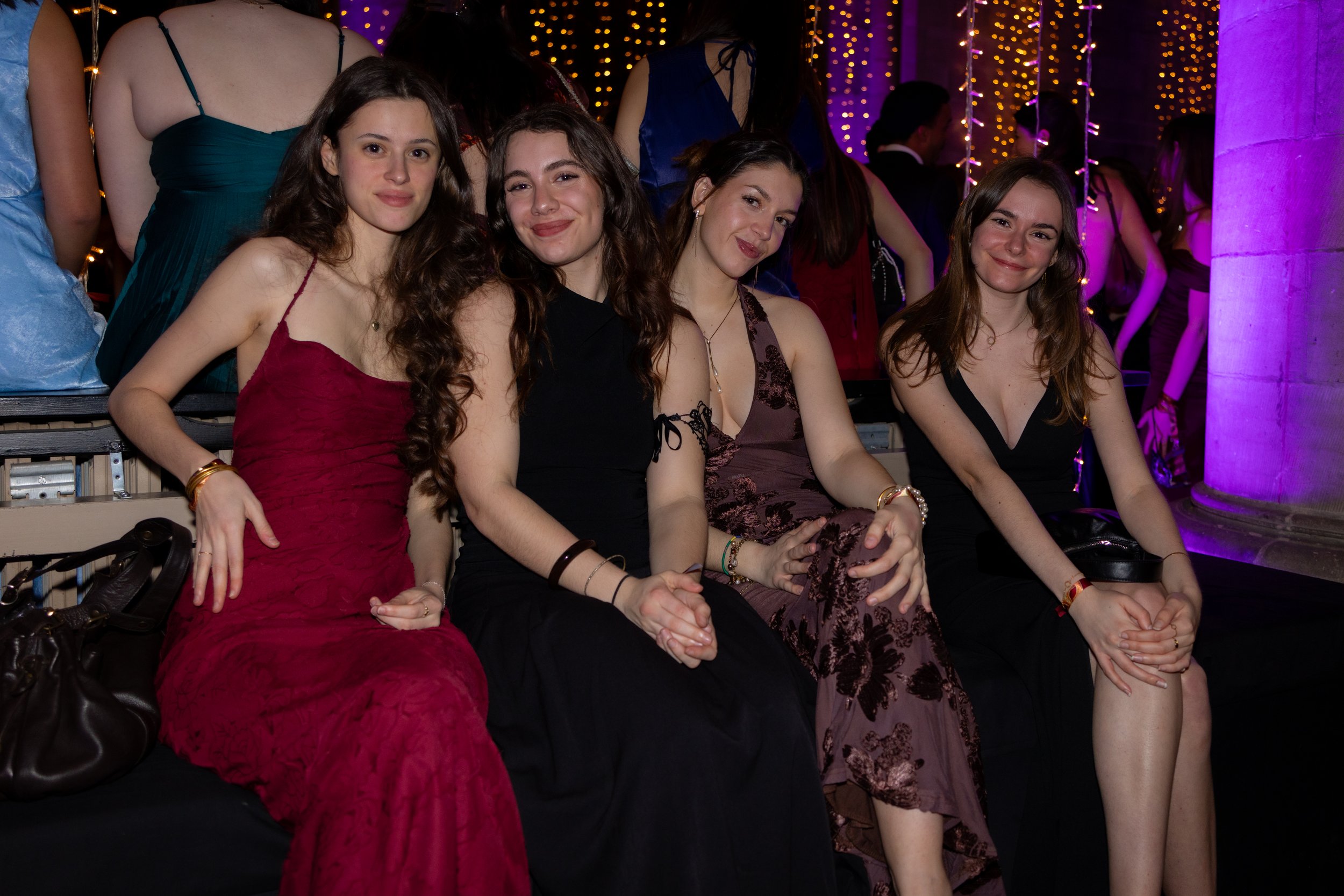 Four young women sitting together at a social event, smiling, with a bar and other guests in the background under decorative string lights.