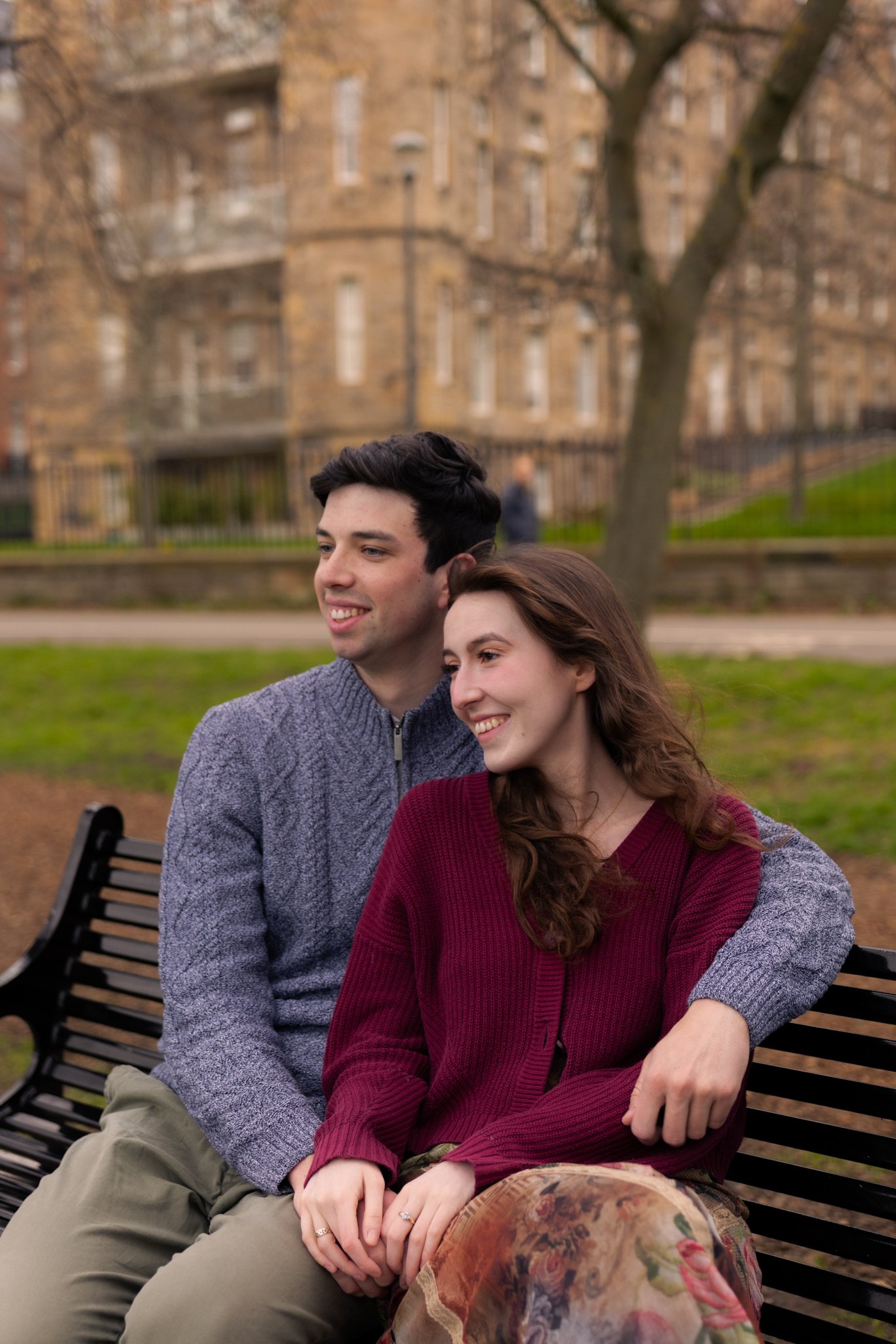 A young couple sitting close on a park bench with their arms intertwined, smiling and looking off into the distance. The background features a large brick apartment building and leafless trees, suggesting fall or winter.