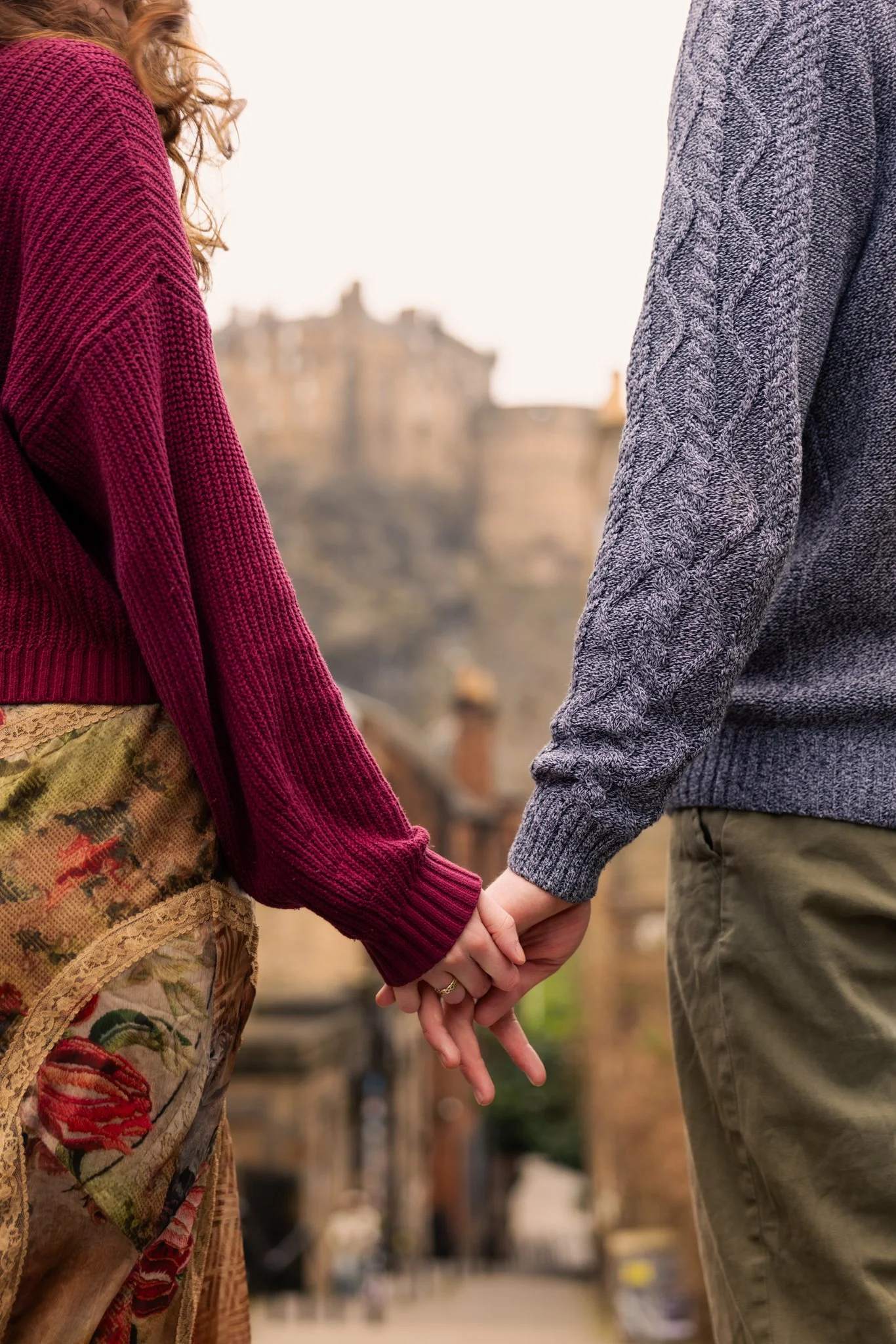 A couple holding hands, standing outdoors, holding hands with their hands intertwined, against a blurred cityscape background.