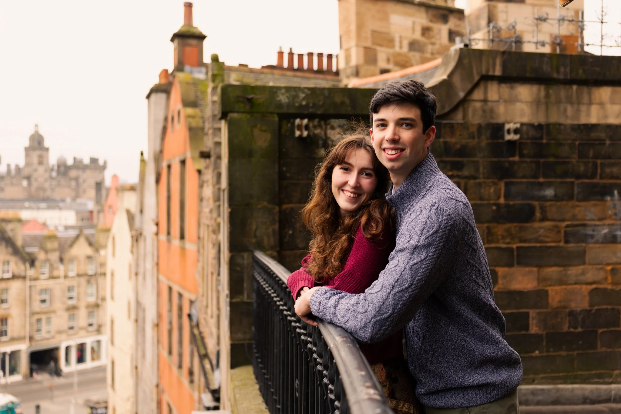 A young couple hugging on a rooftop balcony with city buildings in the background.