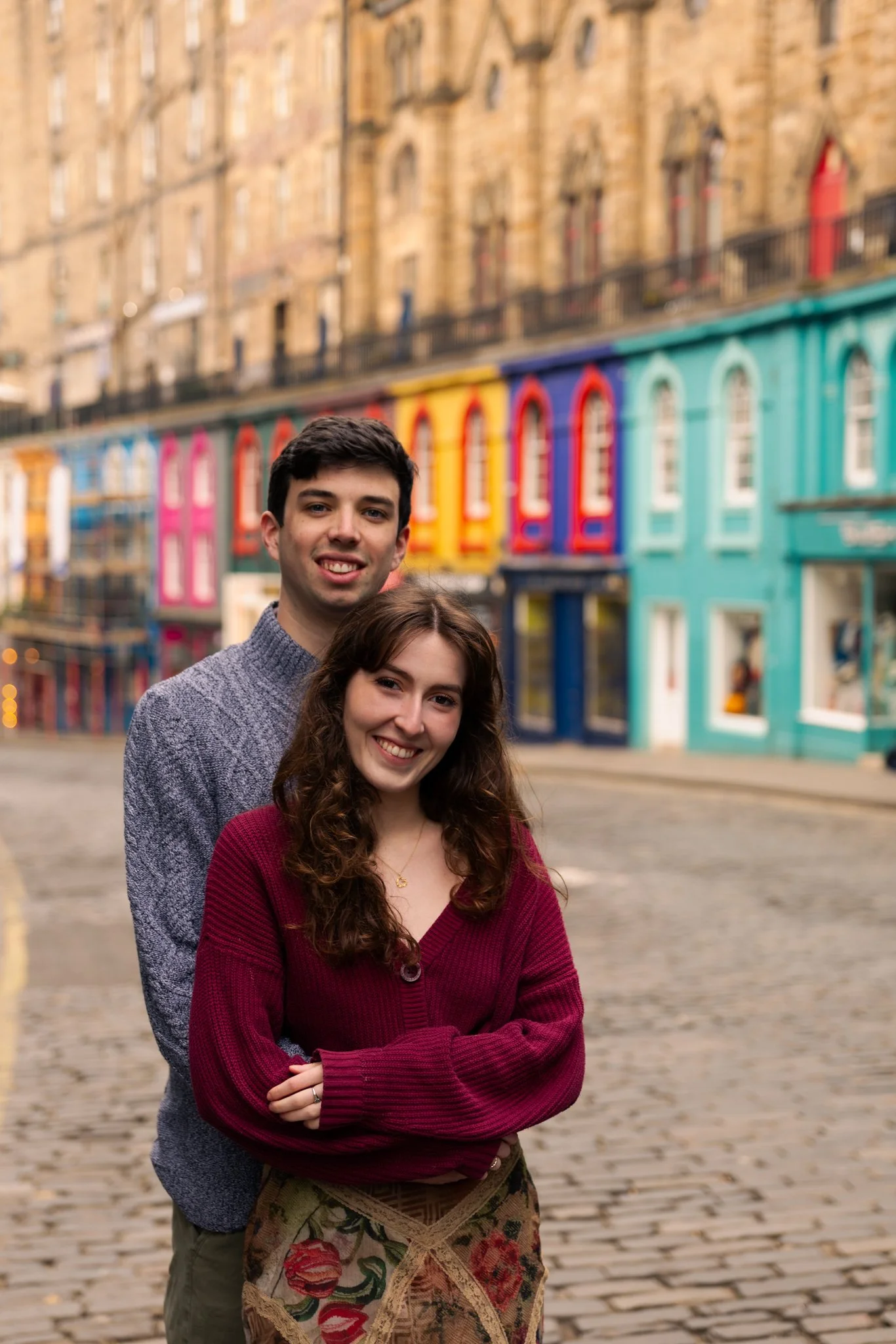 A young man and woman standing on a cobblestone street in front of colorful buildings with windows and a railing on the upper level, in an urban setting.