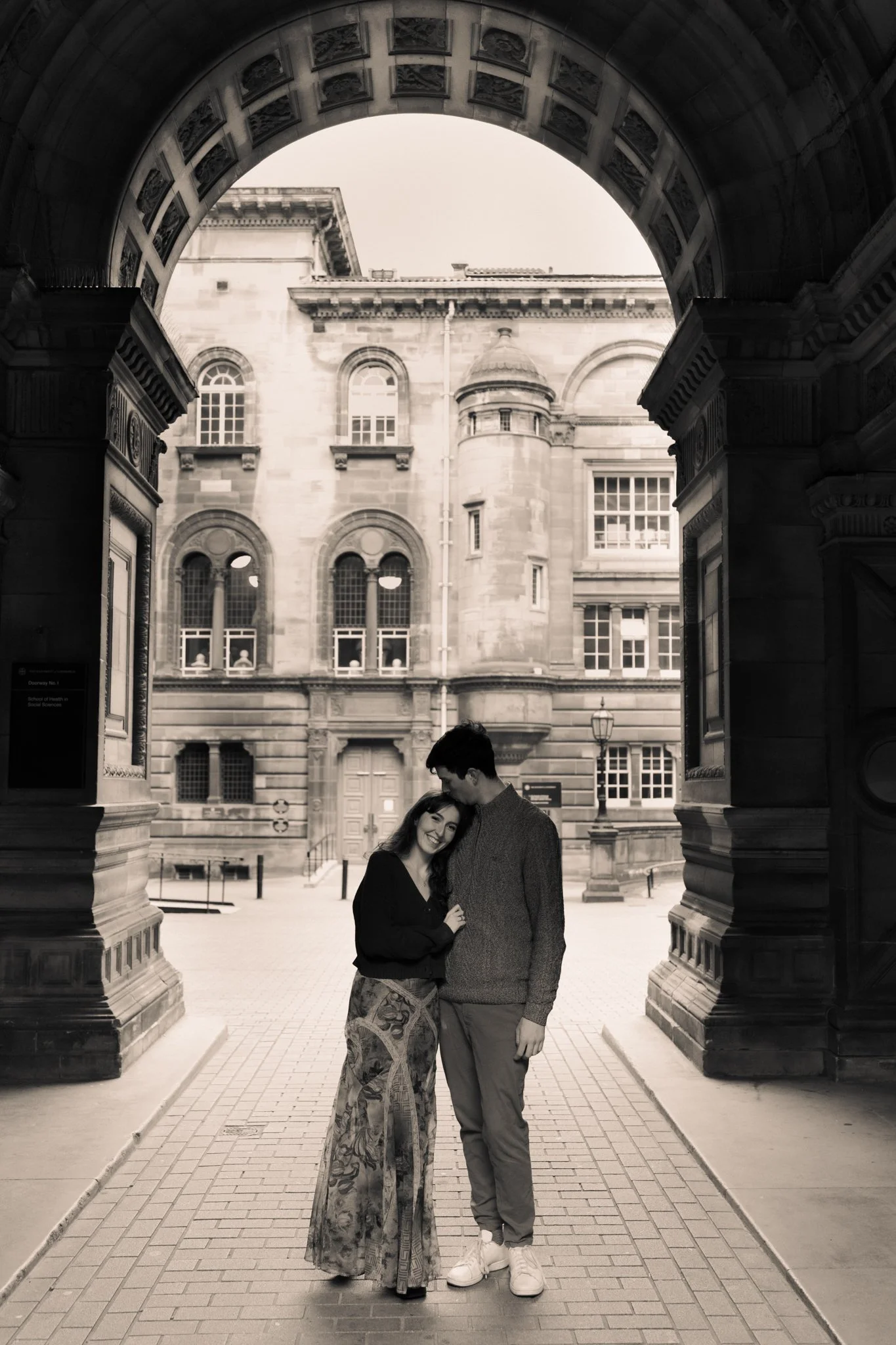 A black and white photo of a couple standing under an ornate archway, with historic buildings in the background. The woman is smiling and leaning into the man, who is looking down at her. They are embracing each other.