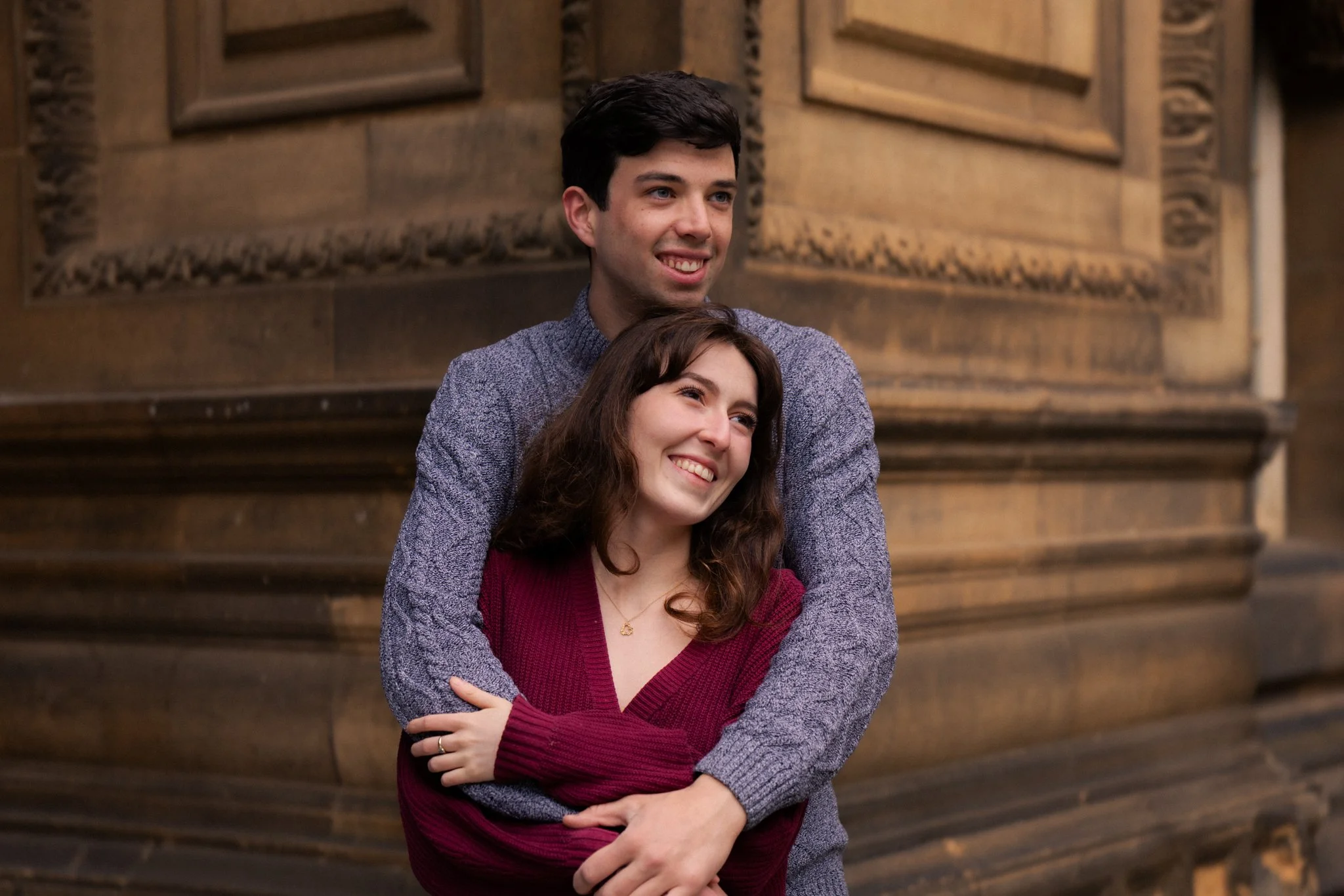 A young man with dark hair wearing a purple sweater is smiling while hugging a young woman with brown hair wearing a maroon sweater. They are standing in front of a wooden staircase.