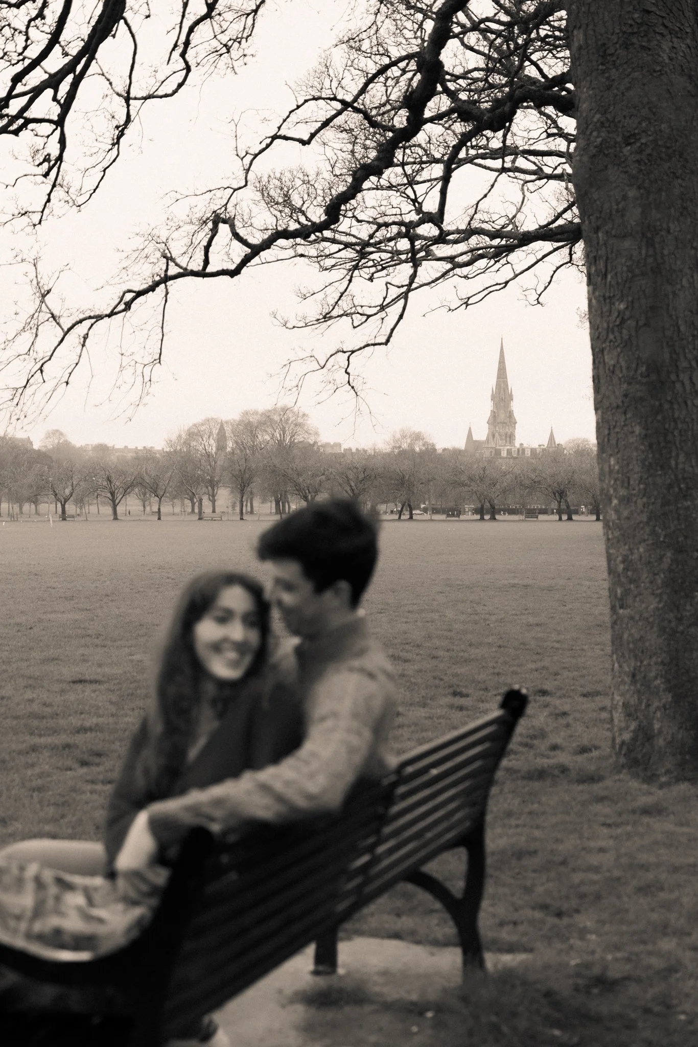 A black and white photo of a park with a woman sitting on a bench in the foreground and a blurred couple beside her. In the background, there are leafless trees and a church steeple.
