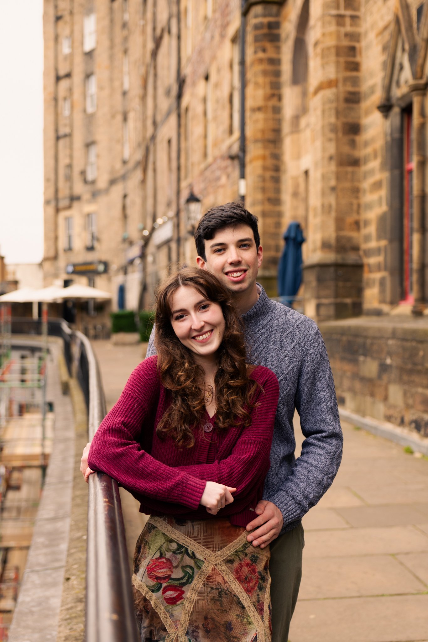A young man and woman smiling standing close together on a bridge in an old city with stone buildings in the background.