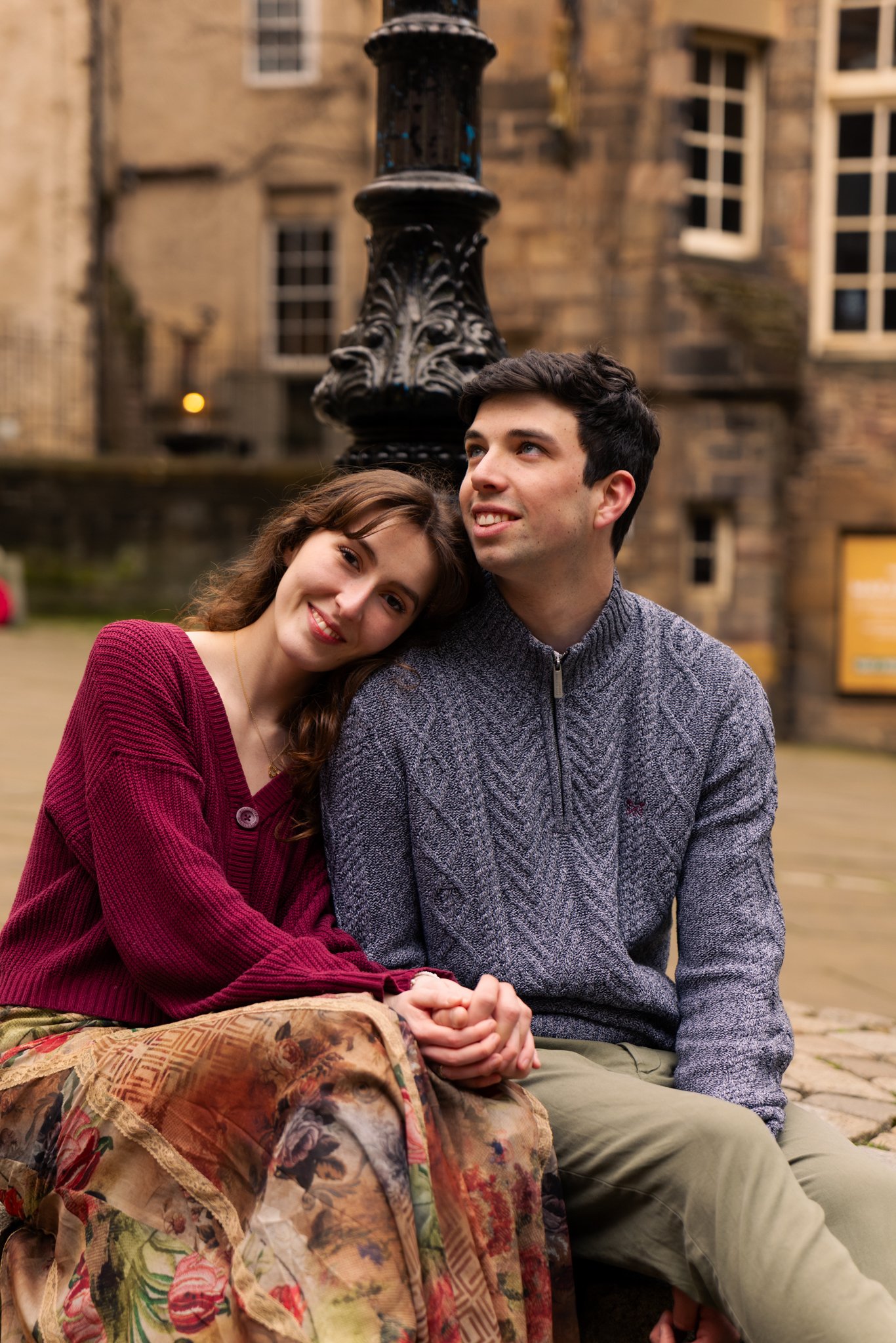 A young man and woman sitting outdoors in front of an old brick building, smiling and leaning on each other, with a decorative black lamp post behind them.