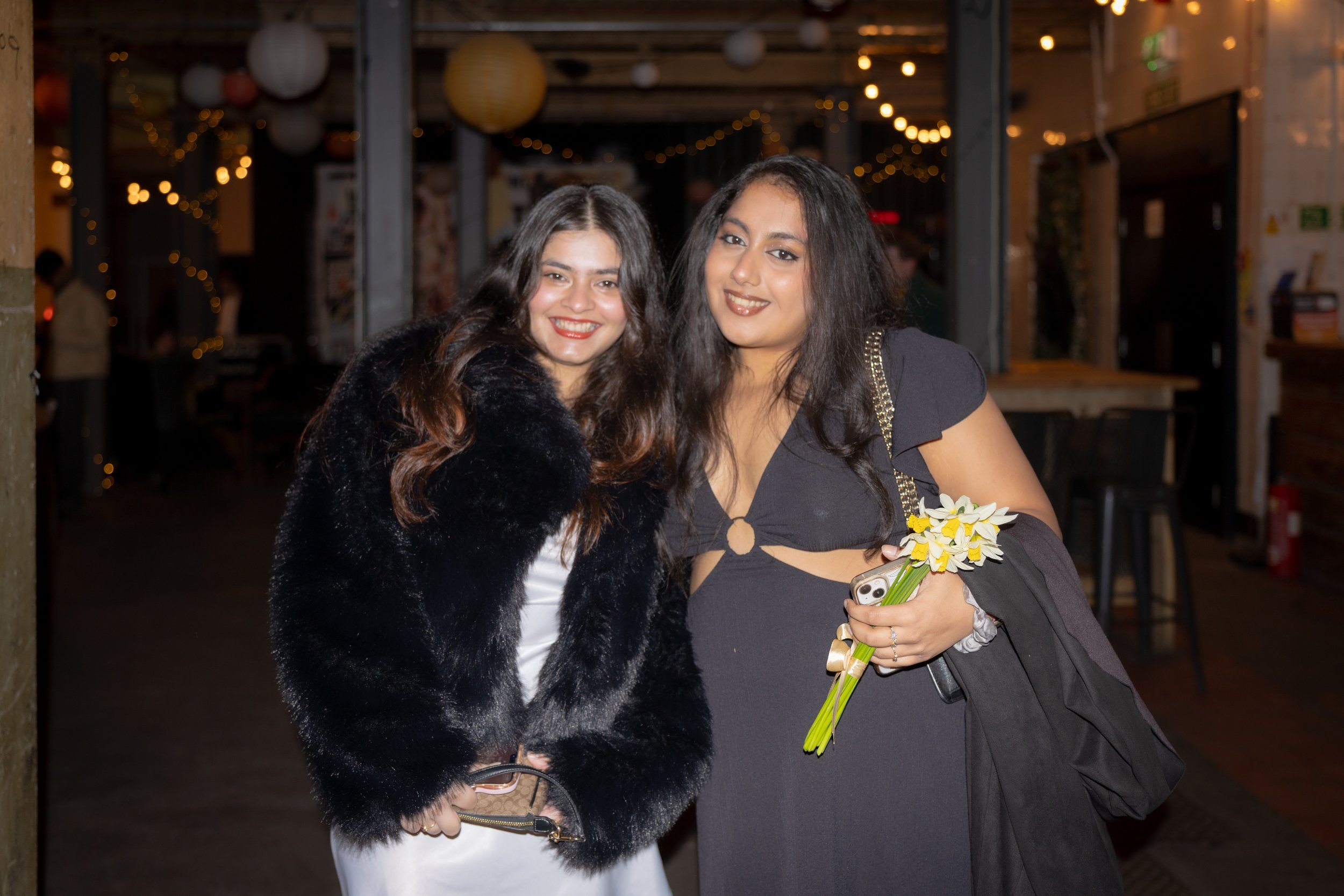 Two women standing close together at night, smiling at the camera, in a decorated venue with string lights and paper lanterns in the background. One is holding a small bouquet of yellow and white flowers, and the other is holding a phone with a strap