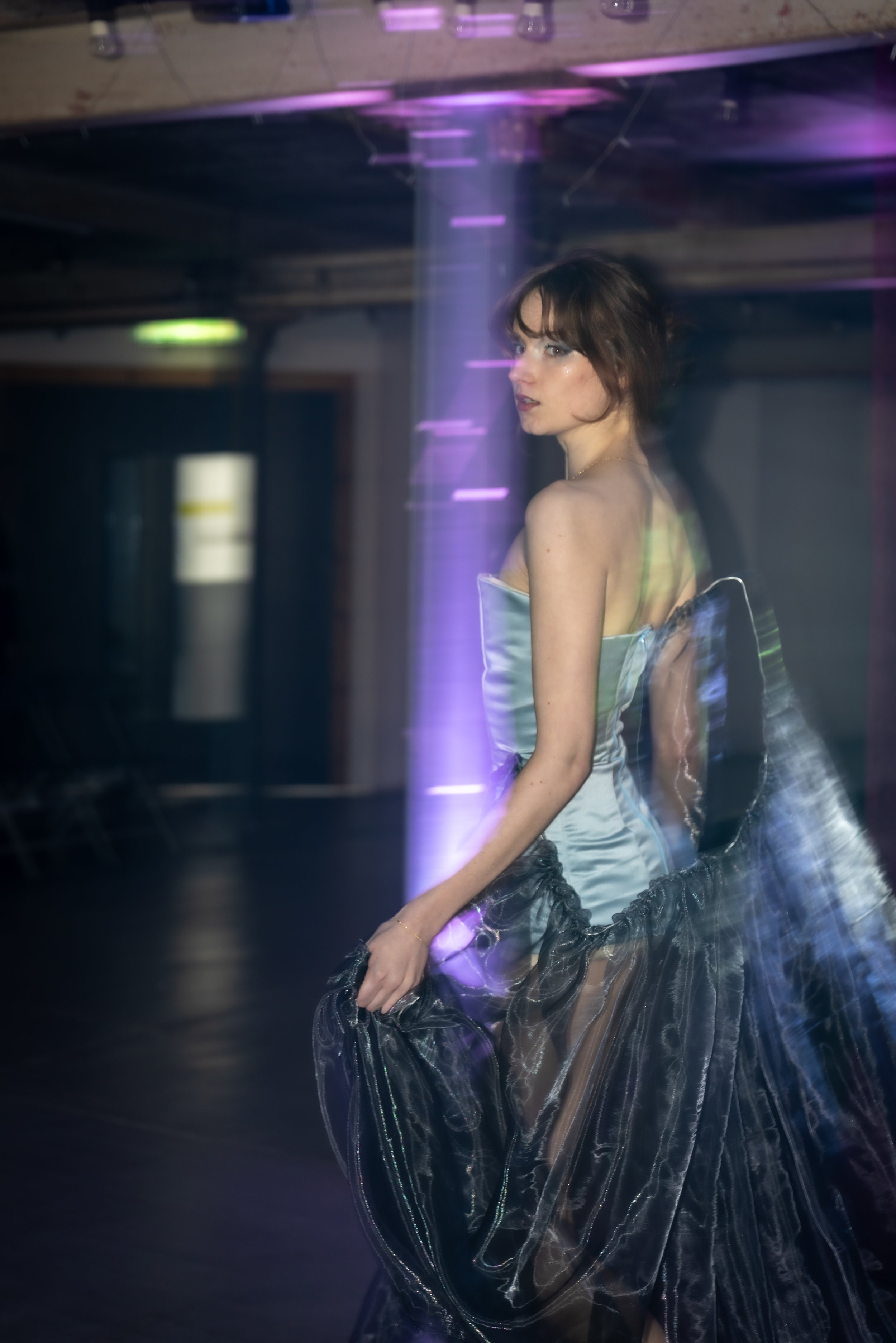 A woman with short brown hair and light makeup standing in a dimly lit space with purple and blue lighting, wearing a strapless satin dress with a ruffled black tulle skirt, holding her right arm with her left hand, and looking over her left shoulder