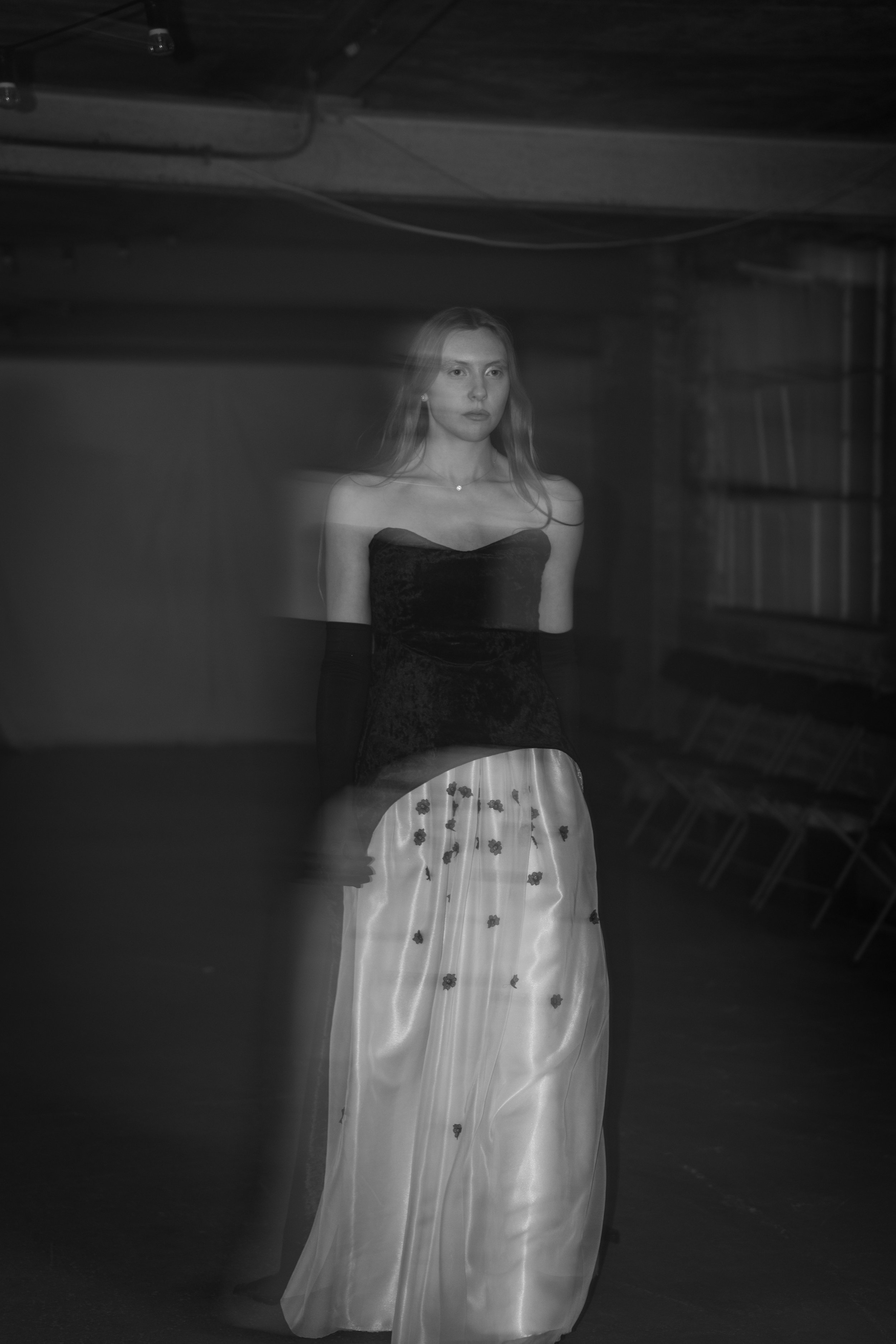 A woman with long hair wearing an off-the-shoulder black dress and a long light-colored skirt with a floral pattern, standing in a dimly lit room.
