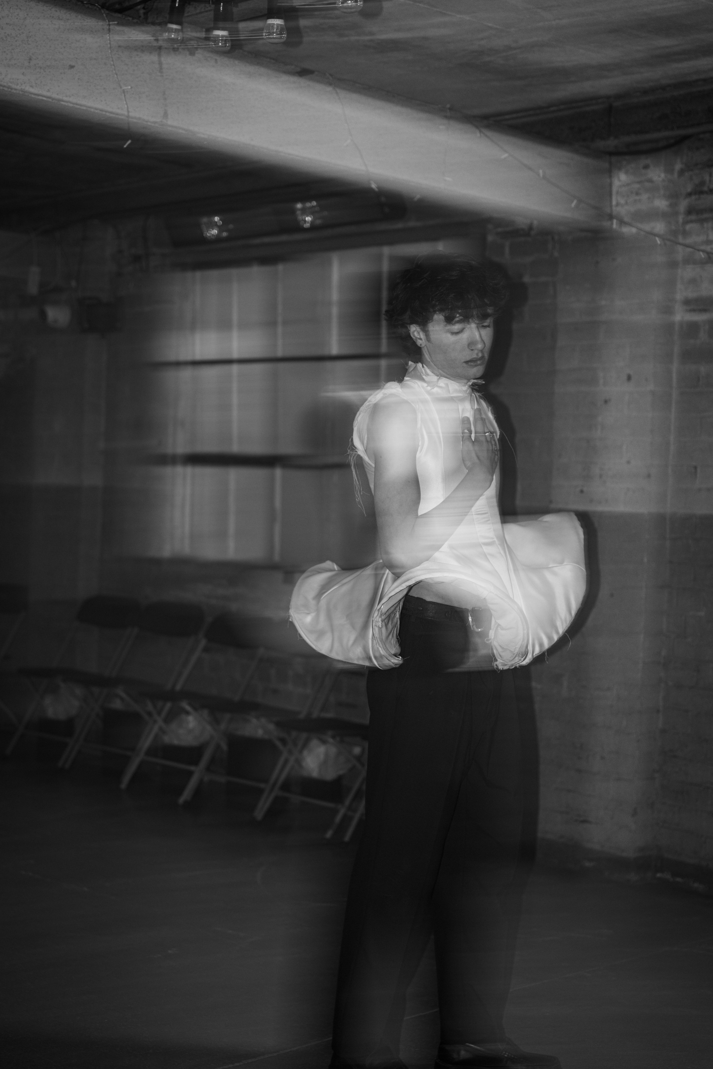 A person with dark, curly hair and light clothing standing in front of a mirror, visible reflections, with a background of a brick wall and chairs.