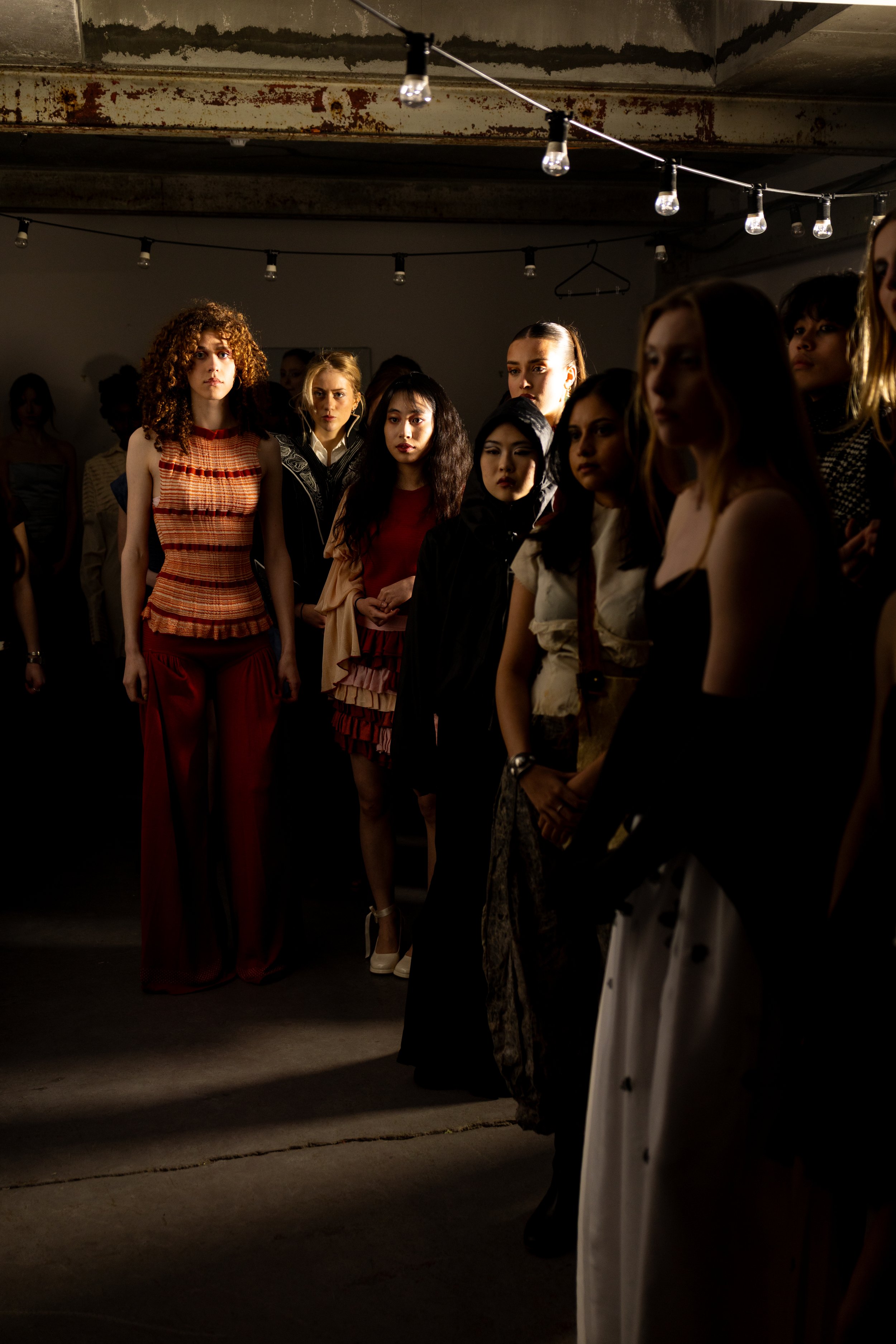 A group of diverse women standing together in a dimly lit, industrial-style room with exposed concrete and rusted metal beams, some with serious expressions.