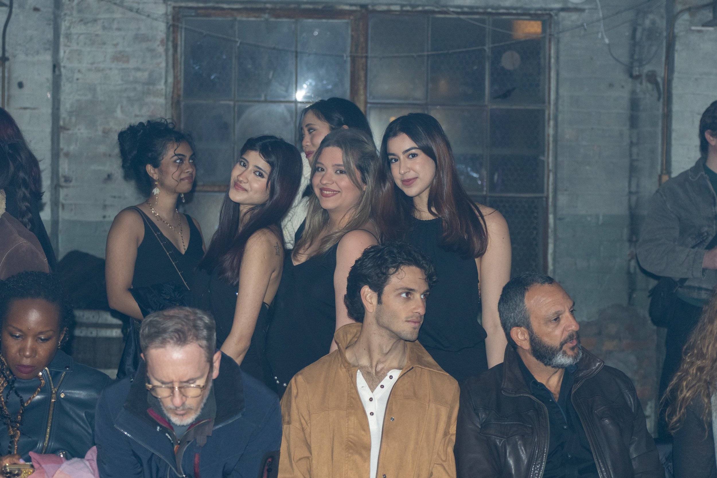 Group of people, mostly women, standing and sitting in a dimly lit industrial-style venue with exposed brick walls and large metal-framed windows, attending an event or gathering.