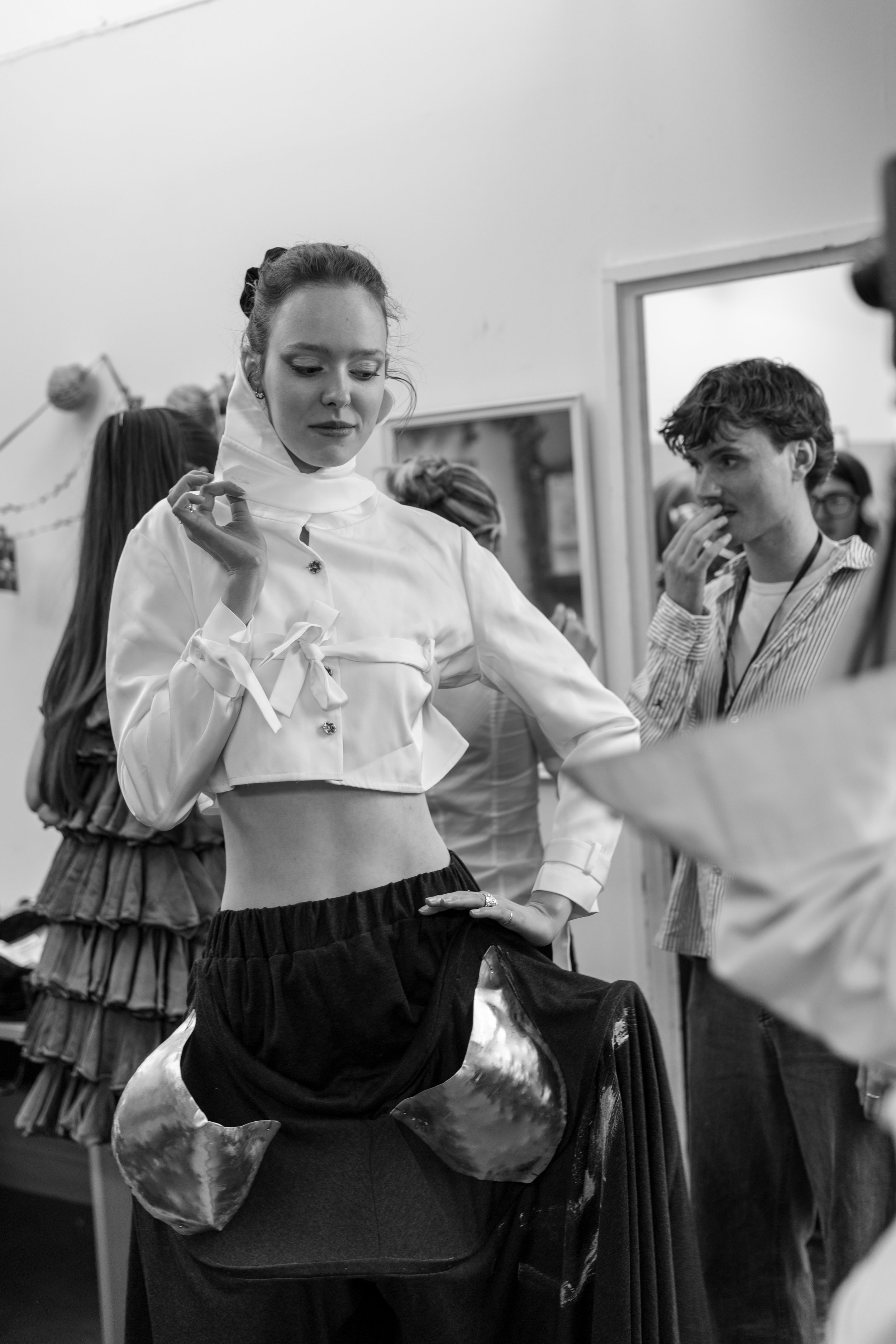 A woman with light skin and dark hair in a bun wears a white blouse with buttons and a black skirt. She is adjusting her blouse, standing next to a person wearing a patterned shirt who appears to be talking to her. In the background, there are other 