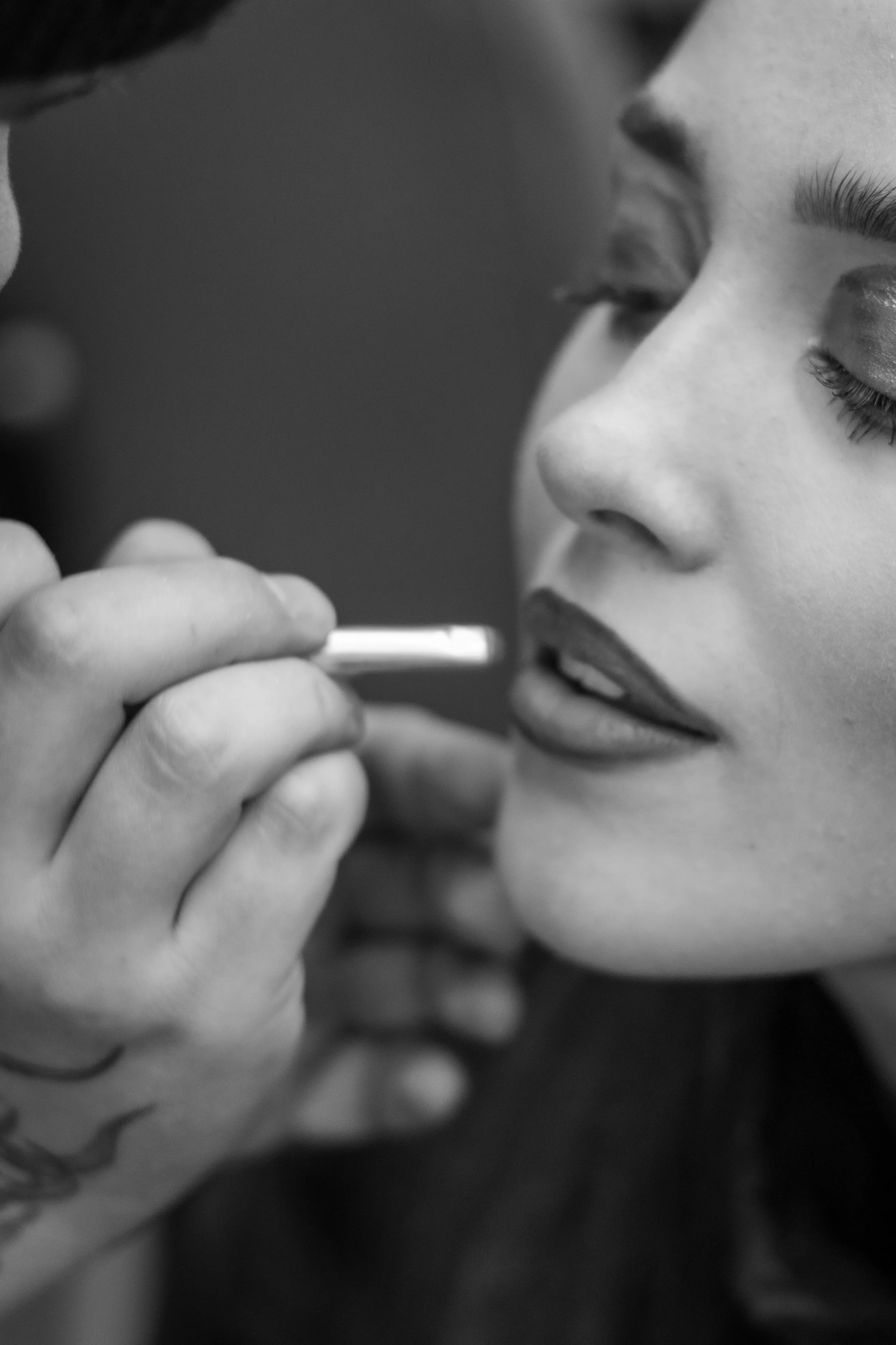 Close-up black and white photo of two people, one with closed eyes, close to each other, with the person on the left holding a small tube near the other's lips, suggesting an intimate or caring moment.