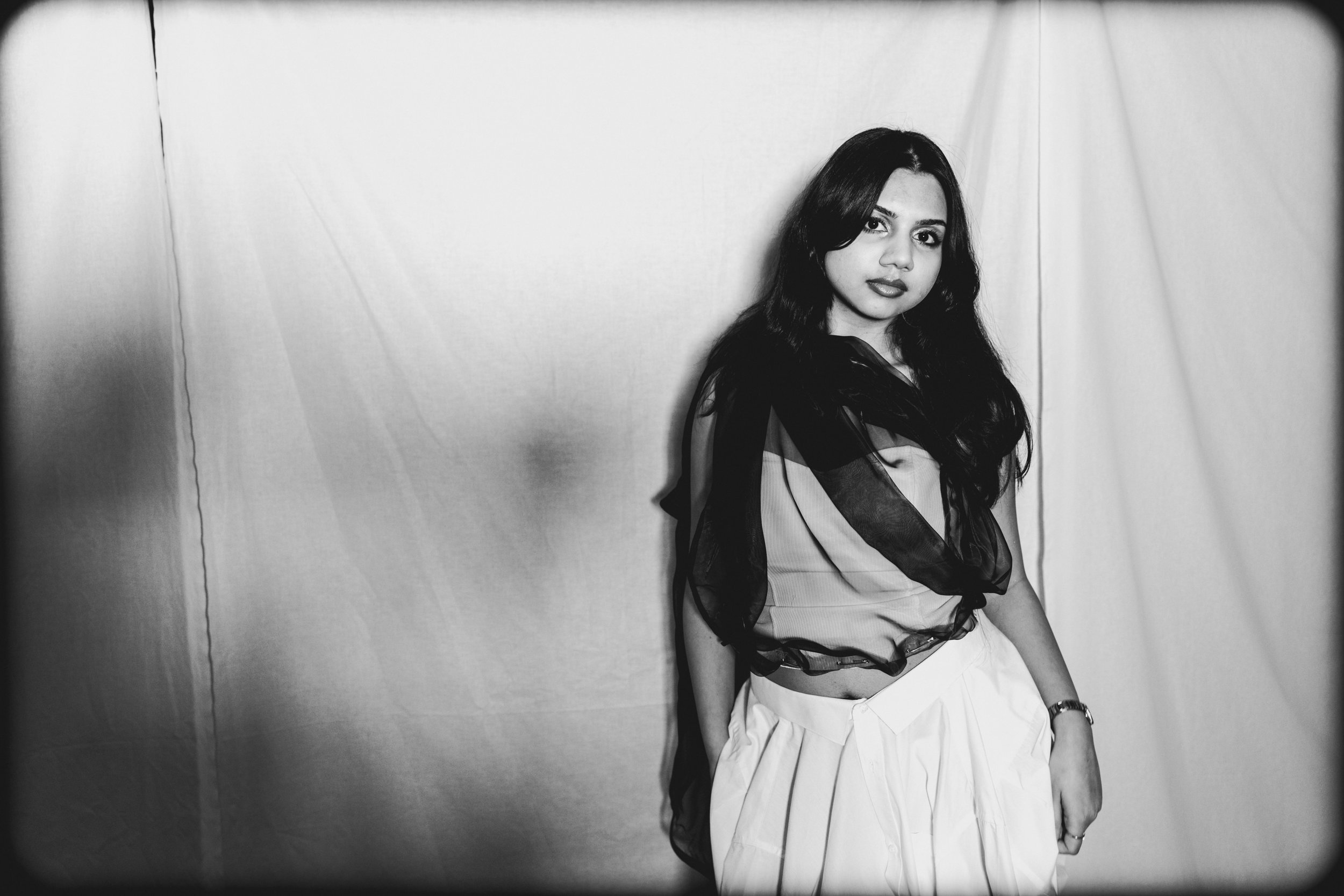 Black and white photo of a woman with long dark hair wearing a dress with sheer black and solid fabric sections, standing against a plain backdrop.