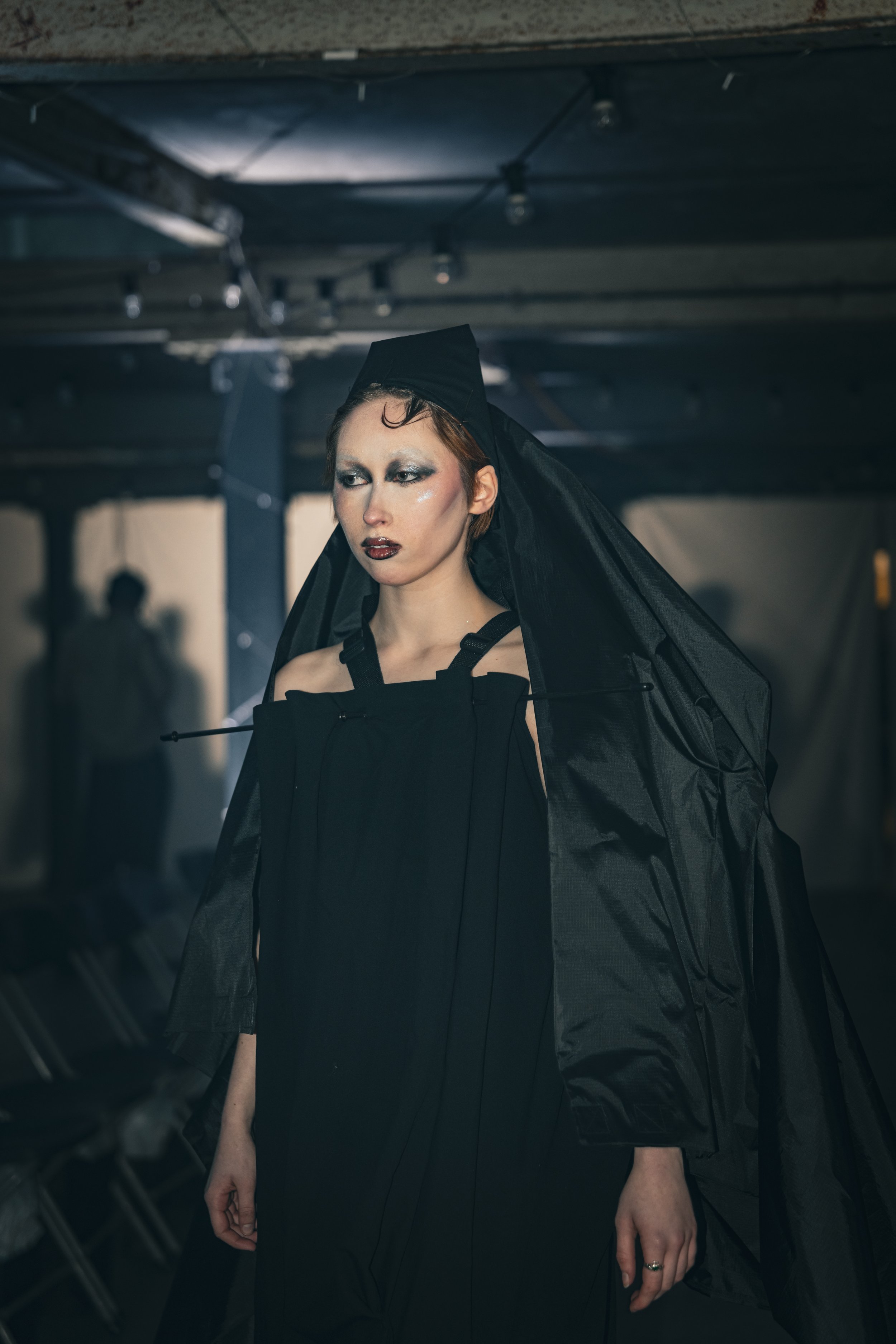 A woman with makeup wearing a black outfit and headpiece, standing in a dark industrial setting with metal beams.