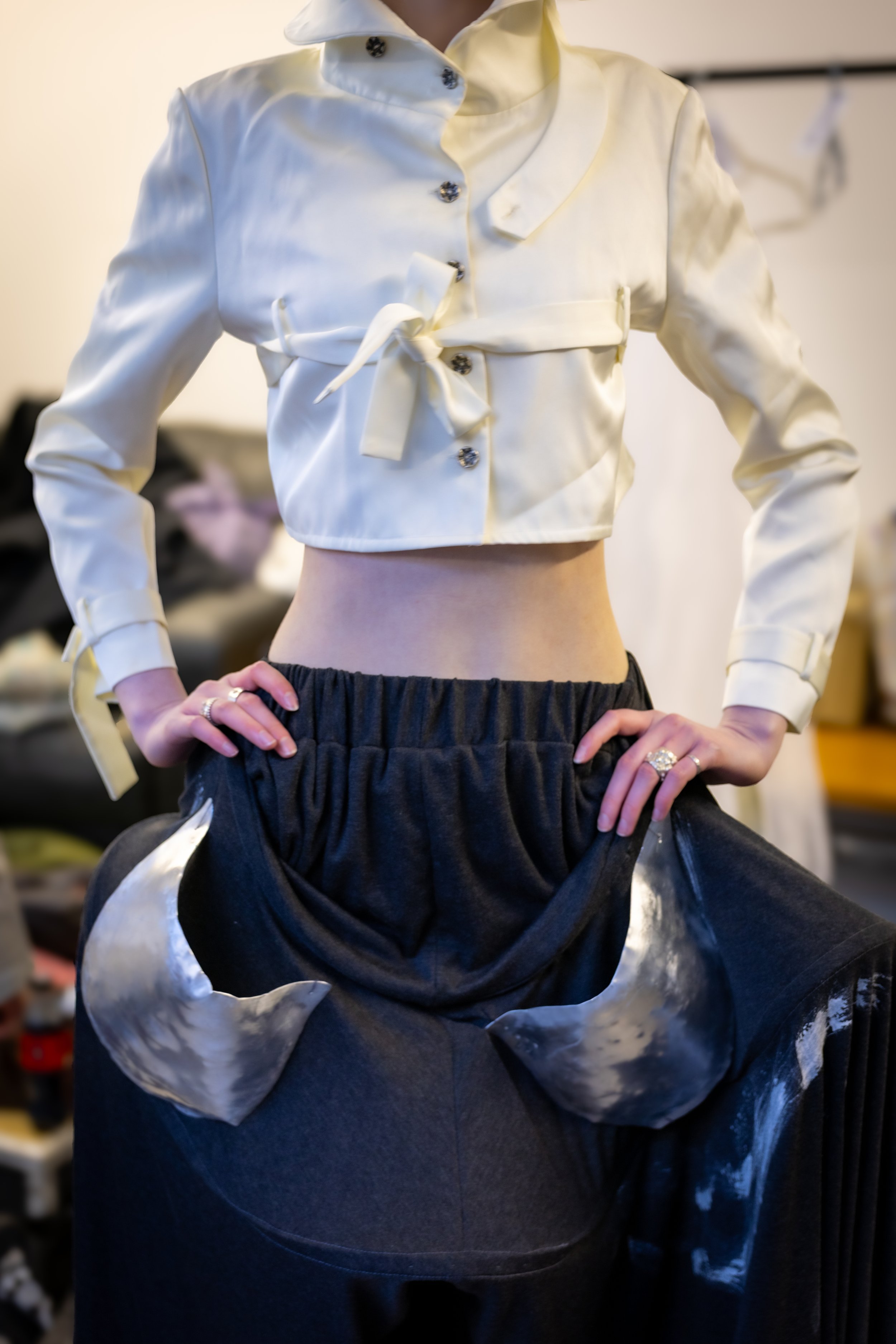 Person wearing a shiny, cream-colored cropped jacket with buttons, a belt, and long sleeves, paired with black, shiny, high-waisted pants.