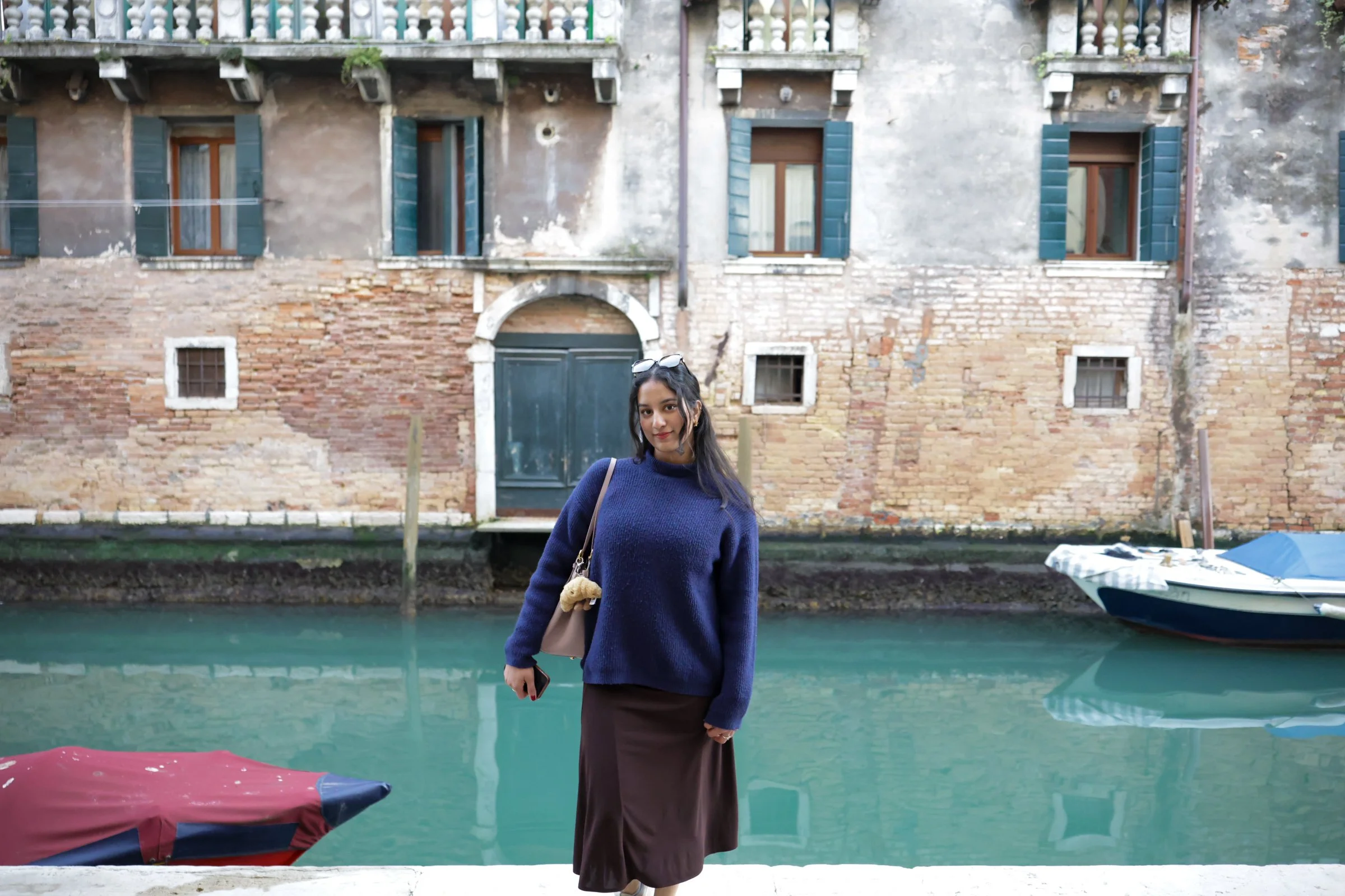 A woman in a navy sweater and brown skirt standing by a canal with boats and an old building with blue shutters in the background.
