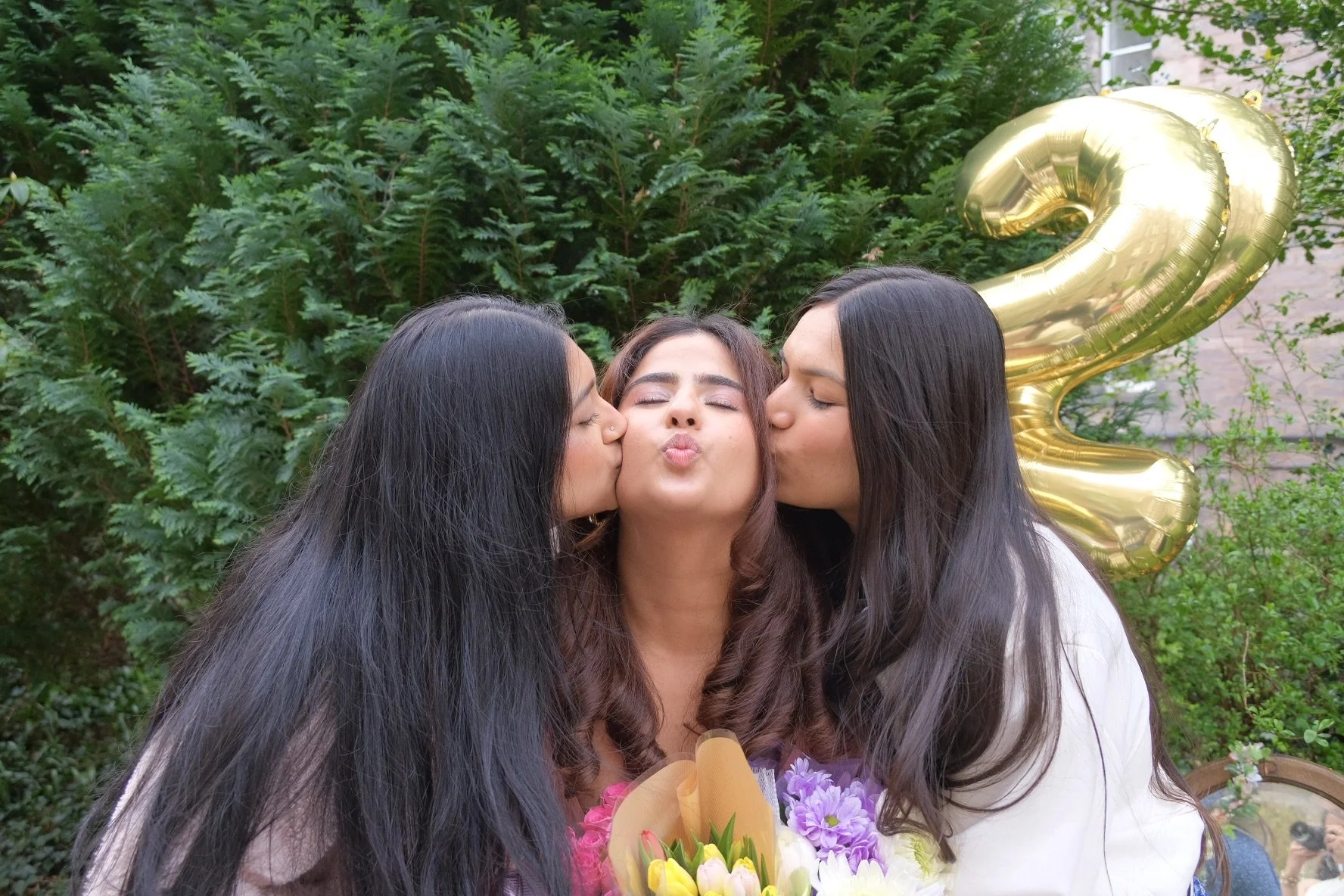 Three women celebrating a birthday outdoors, with two women kissing a woman in the middle on each cheek. The woman in the middle is holding a bouquet of flowers, and a large gold balloon shaped as the number 2 is visible behind them.