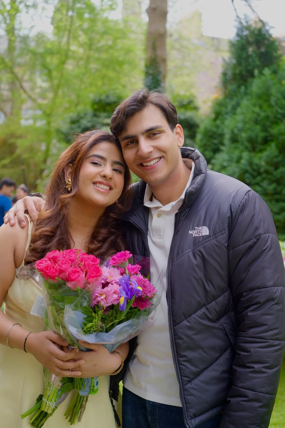 A happy young woman and man hugging outdoors, with the woman holding a bouquet of pink and purple flowers, surrounded by green trees in a park or garden.