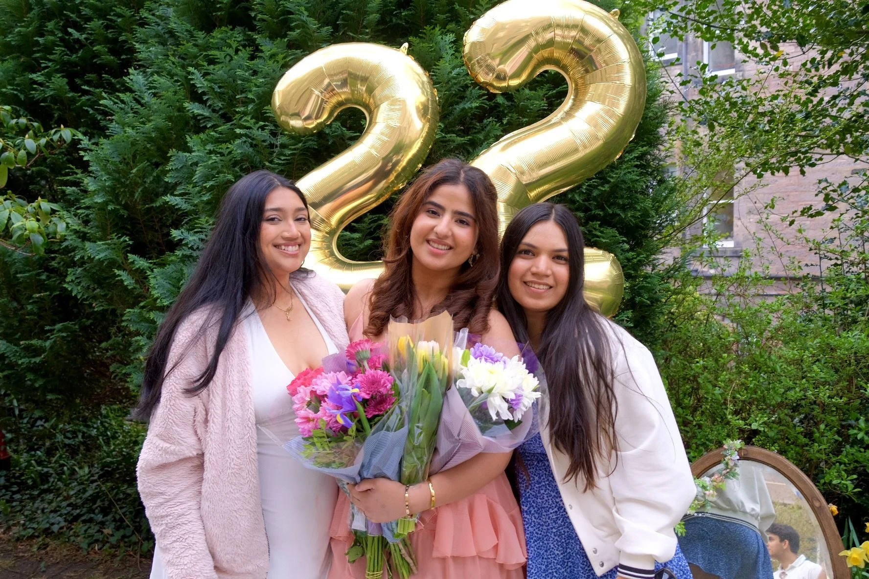 Three young women celebrating graduation outdoors, with large gold balloons forming the number 22, holding bouquets of flowers, surrounded by greenery and a brick building in the background.