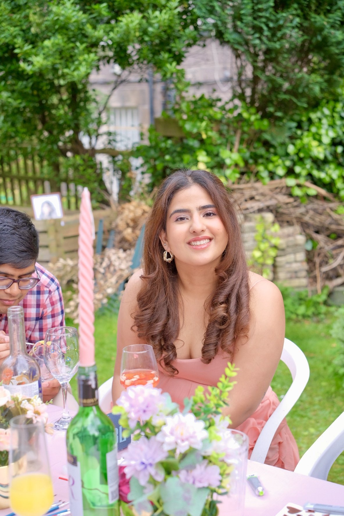 A young woman with wavy brown hair smiling at an outdoor gathering, sitting at a table decorated with flowers, drinks, and bottles.