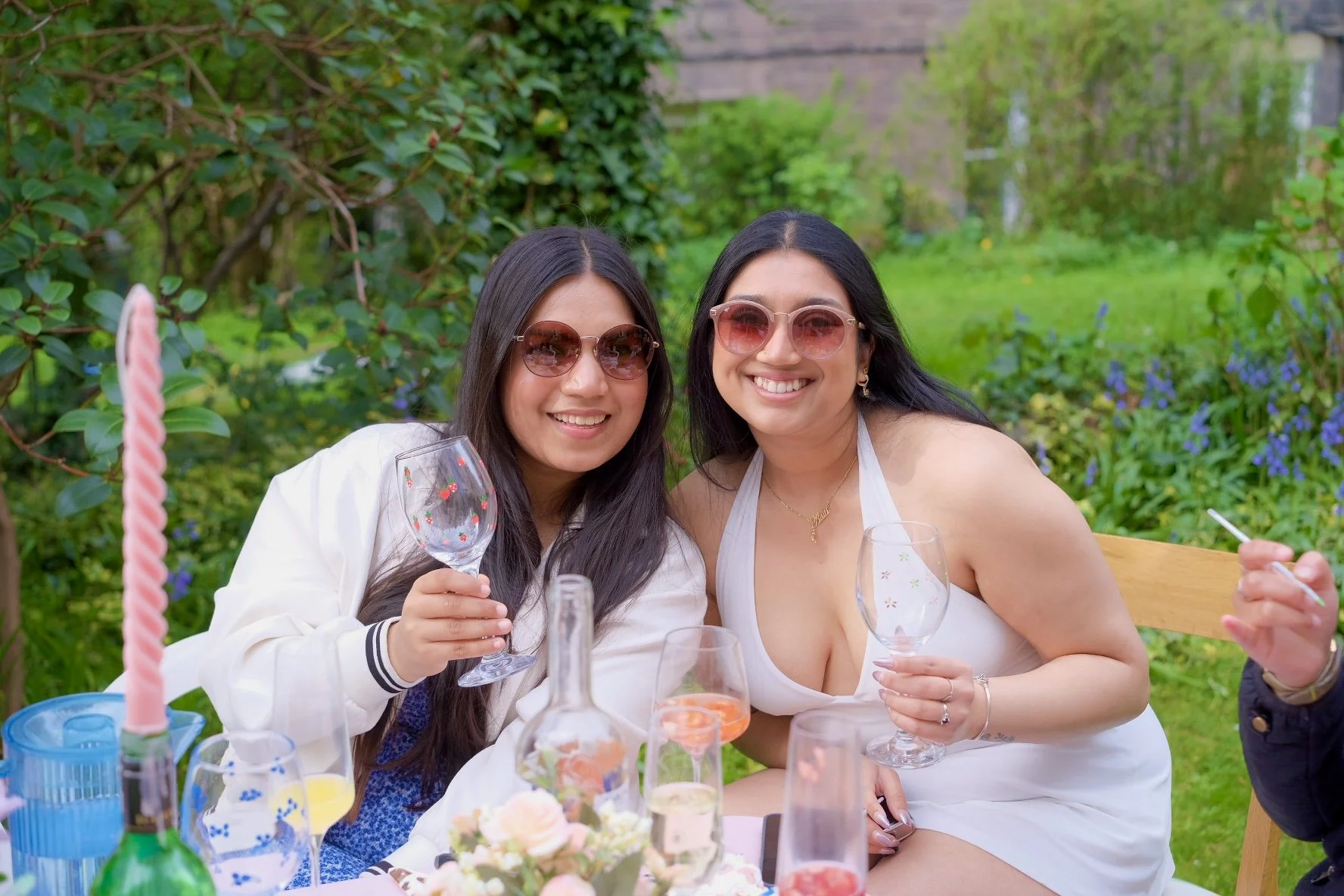 Two women with long dark hair and sunglasses, smiling and holding empty wine glasses, sitting at a table outdoors with a pink birthday candle, bottles, and glasses, surrounded by green foliage and flowers.