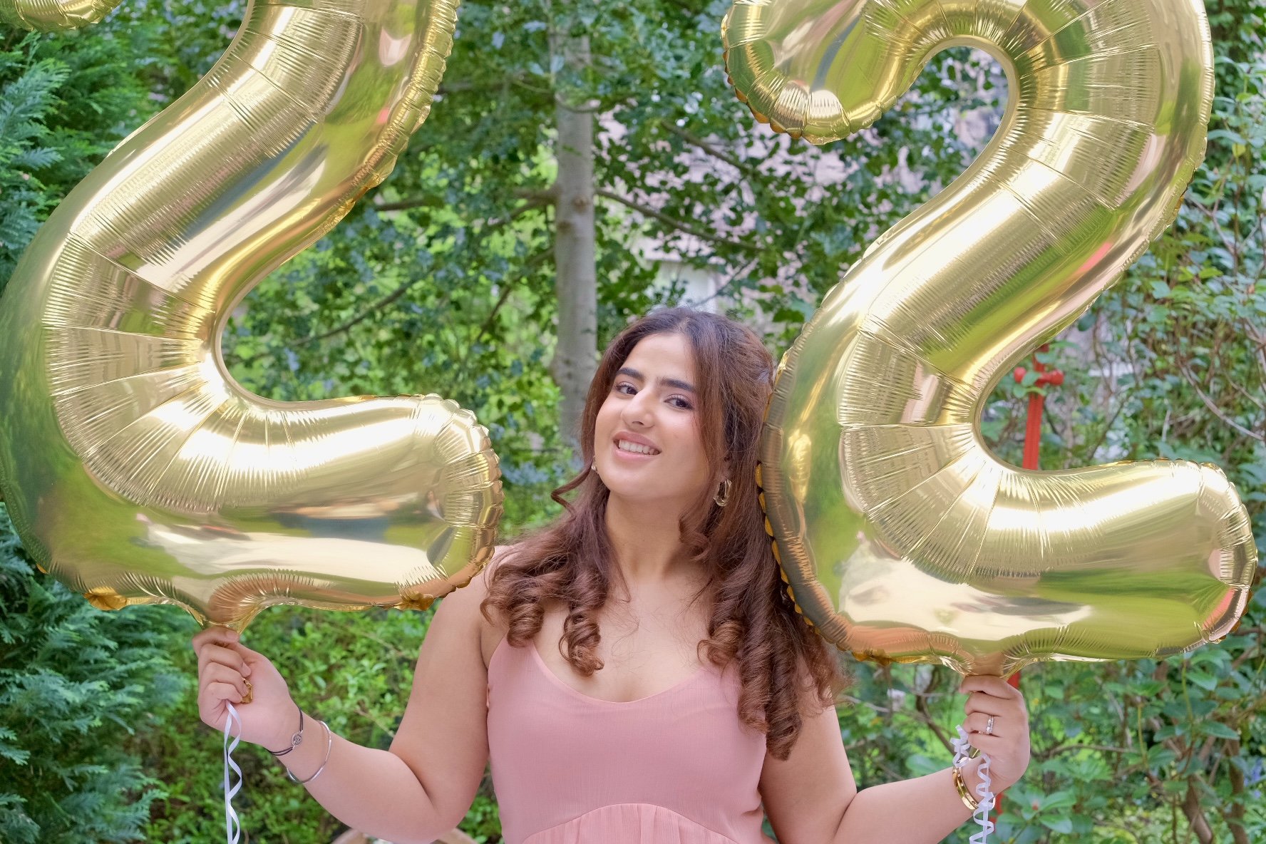 A woman holding two large gold balloons shaped like the number 2 and 3, standing outdoors with green trees in the background for a celebration of her 23rd birthday.