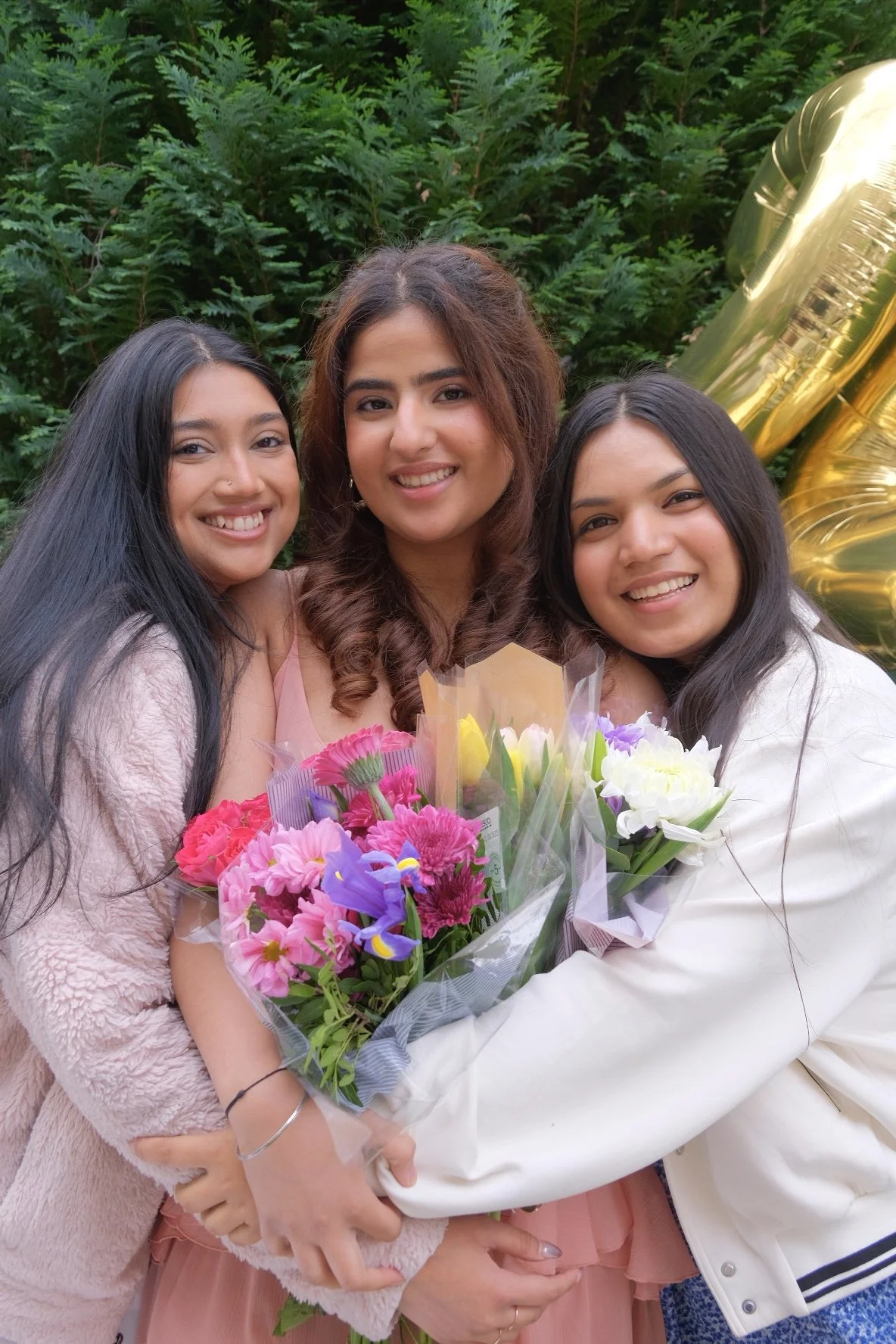 Three young women smiling and holding bouquets of colorful flowers, standing outdoors in front of green foliage and gold balloons.