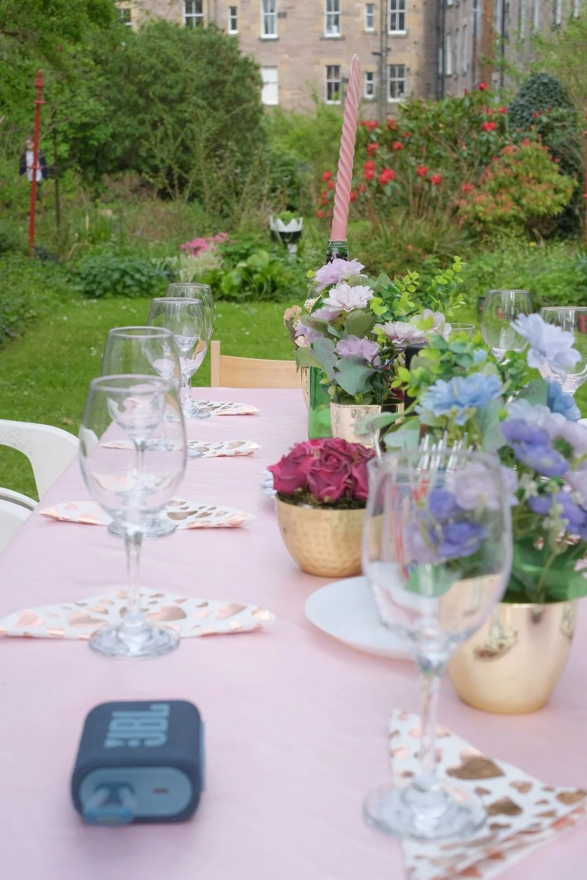 Outdoor table set for a celebration, decorated with pink tablecloth, floral centerpieces with pastel pink, blue, and purple flowers, gold pots, pink candle, and glassware, with a garden and building in the background.