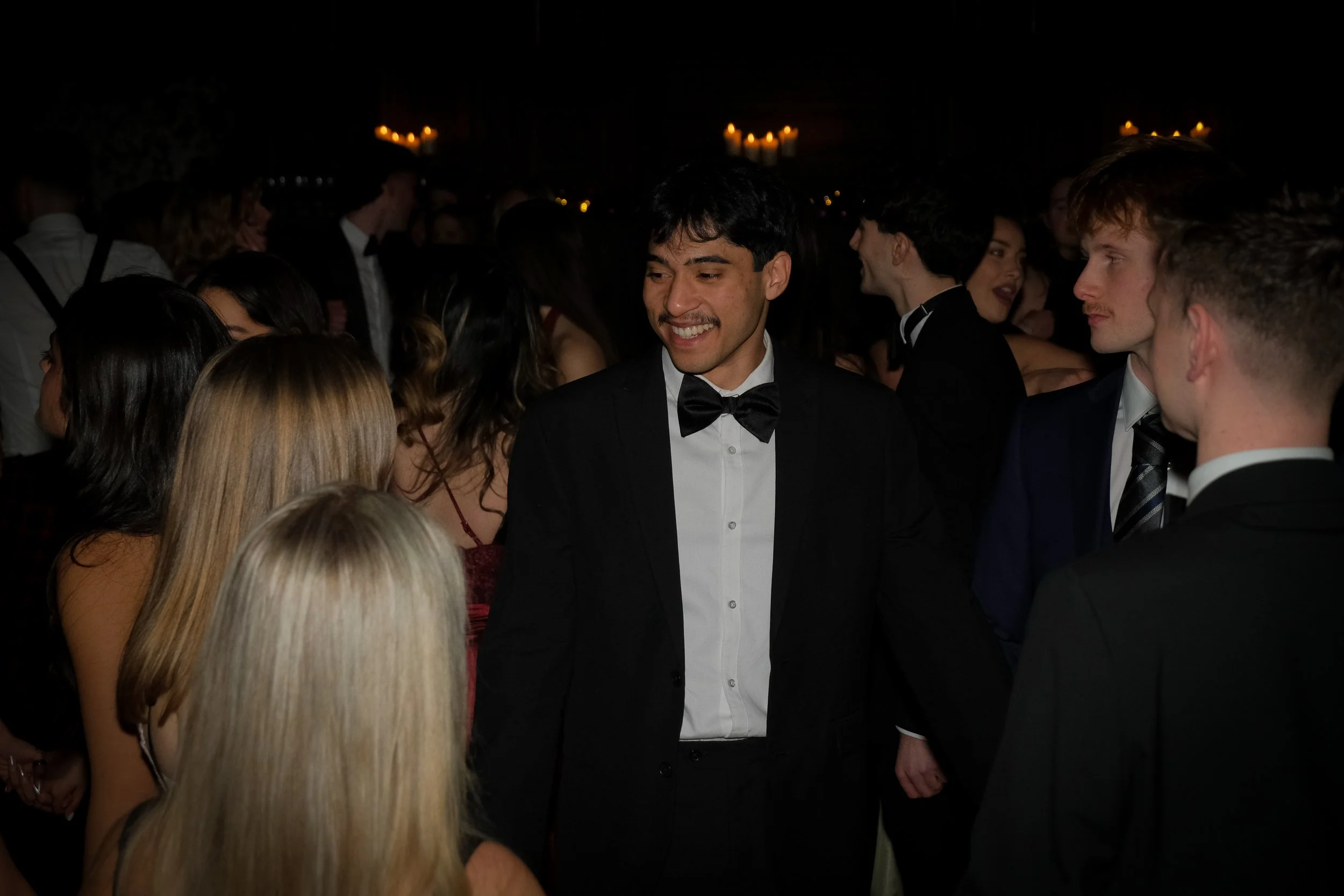 Group of young adults at a formal event, dressed in tuxedos and evening gowns, socializing and smiling.