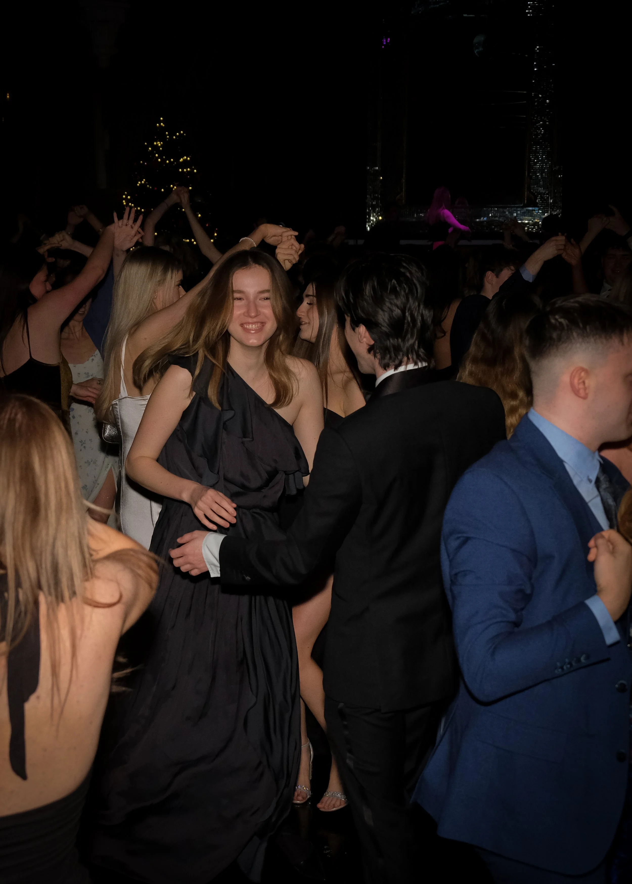 People dancing and socializing at a lively party or celebration in a dark venue with a decorated Christmas tree and city skyline in the background.