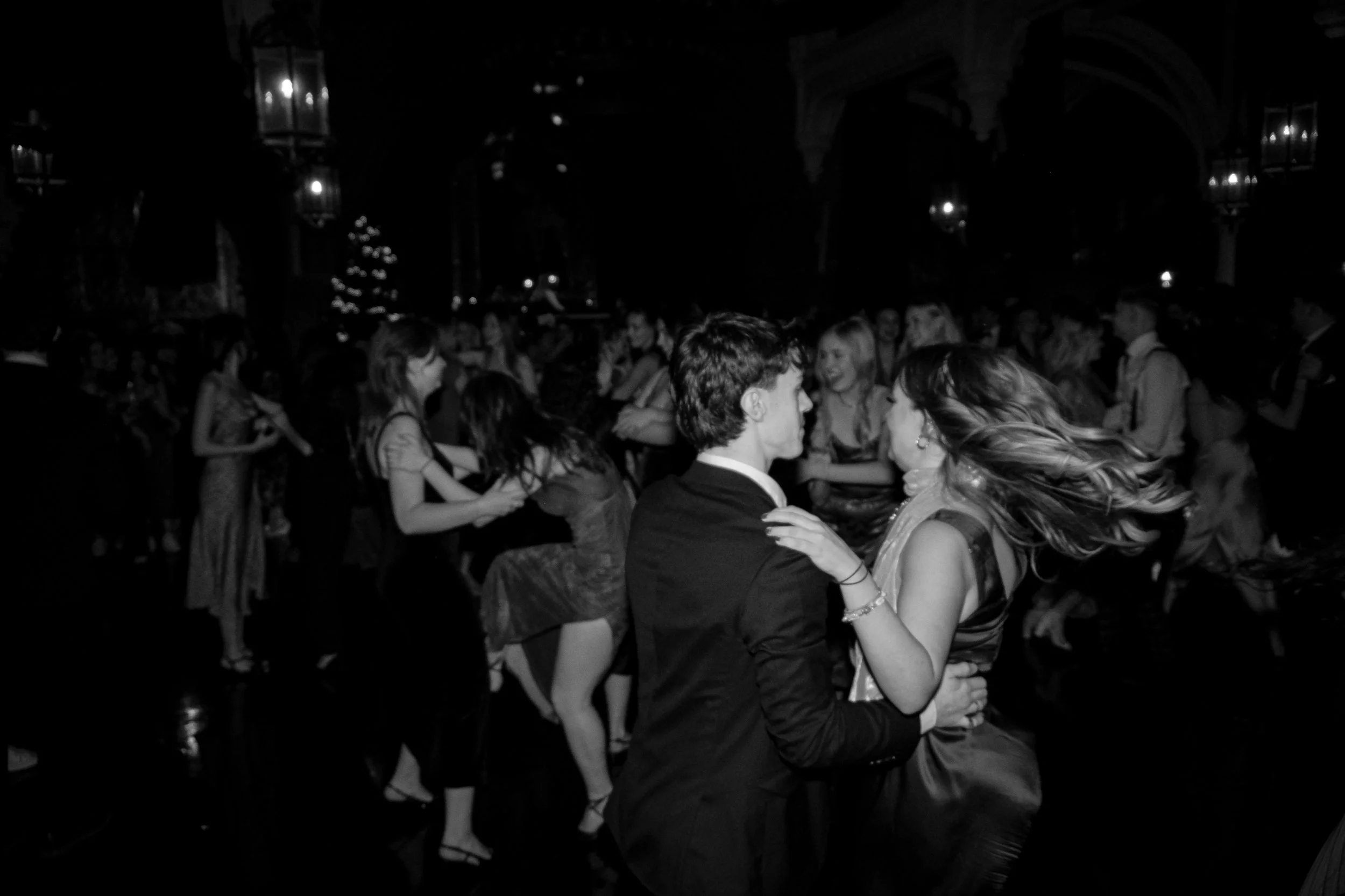 A black and white photo of a couple dancing closely on a crowded dance floor at a formal event or party, with other dancing guests in the background.