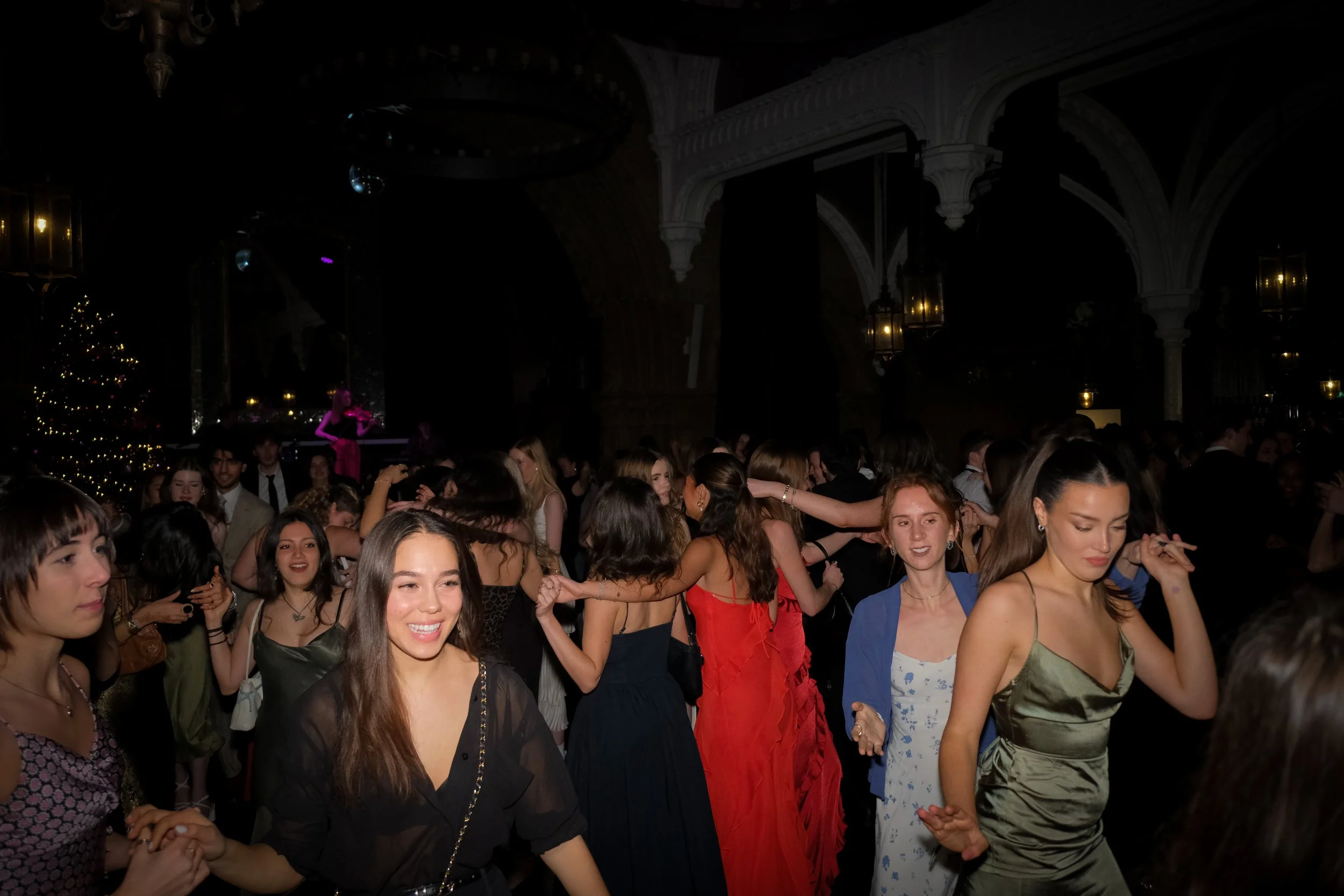 People dancing and socializing at a lively indoor party or dance event with Christmas decorations, including a Christmas tree, in a grand hall with ornate architecture.