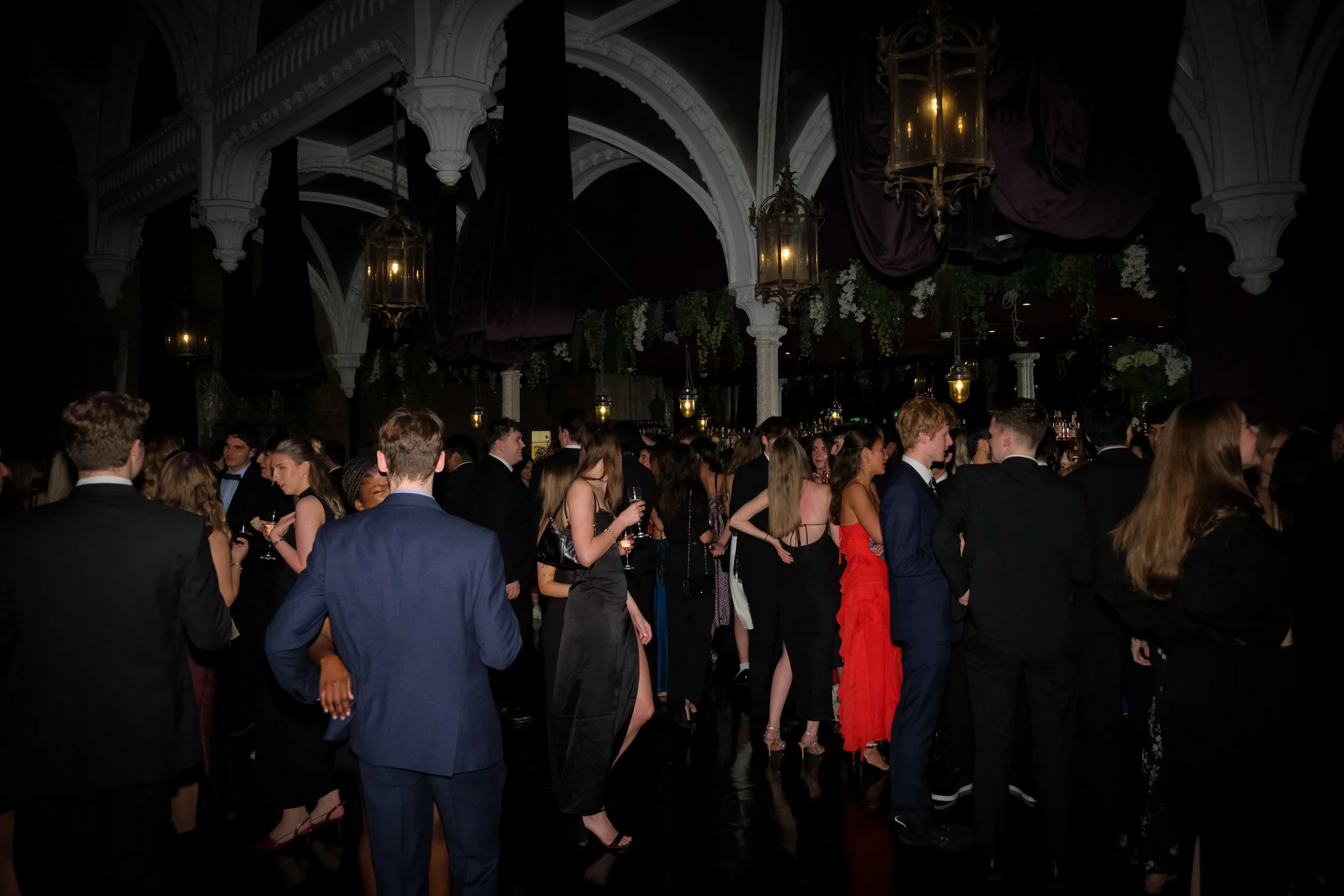 A crowd of elegantly dressed people socializing at a formal event in a dimly lit, ornate venue with chandeliers and floral decorations.