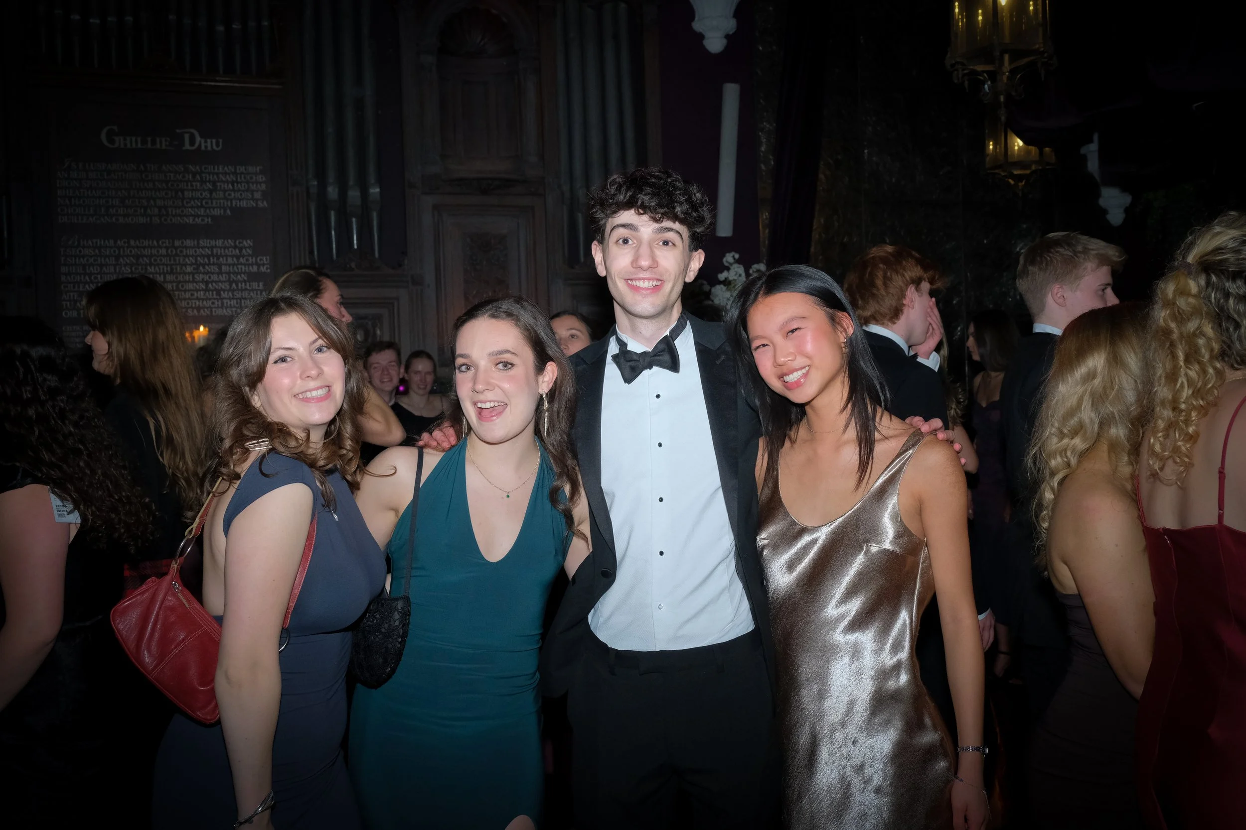 Four young adults dressed in formal attire smiling at a party or formal event, standing close together with arms around each other.