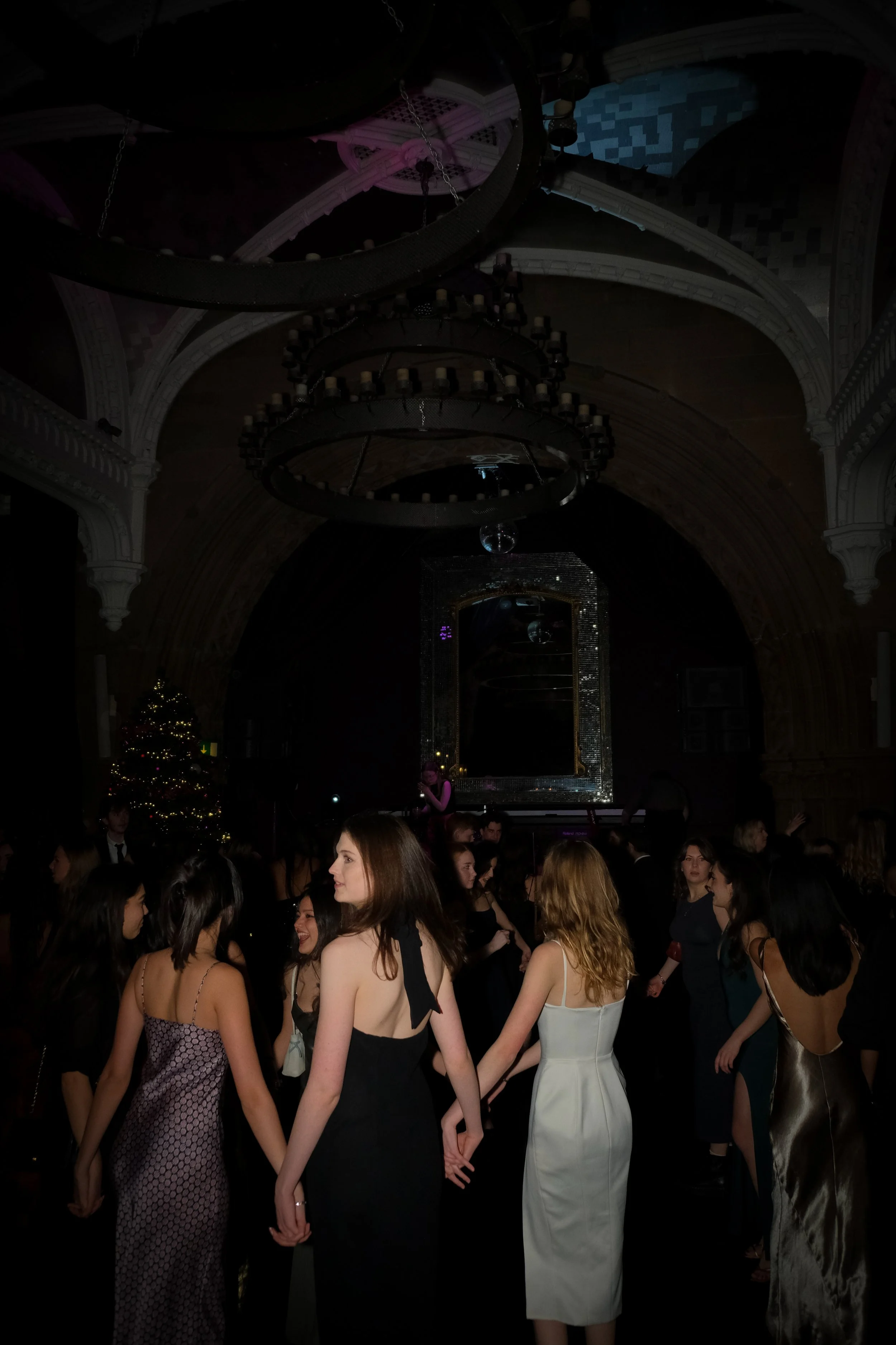 People dancing in a dimly lit ballroom with chandeliers, a Christmas tree, and a large mirror on the wall.