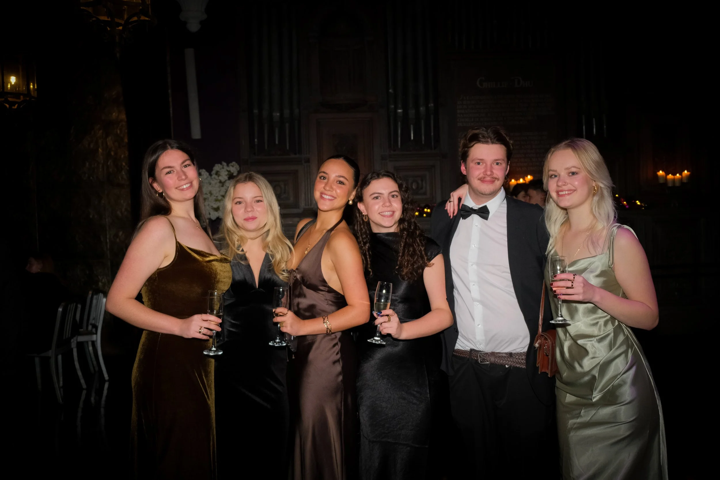Group of seven young people dressed in formal attire, enjoying a celebration and holding glasses of wine or champagne, in an elegant indoor setting