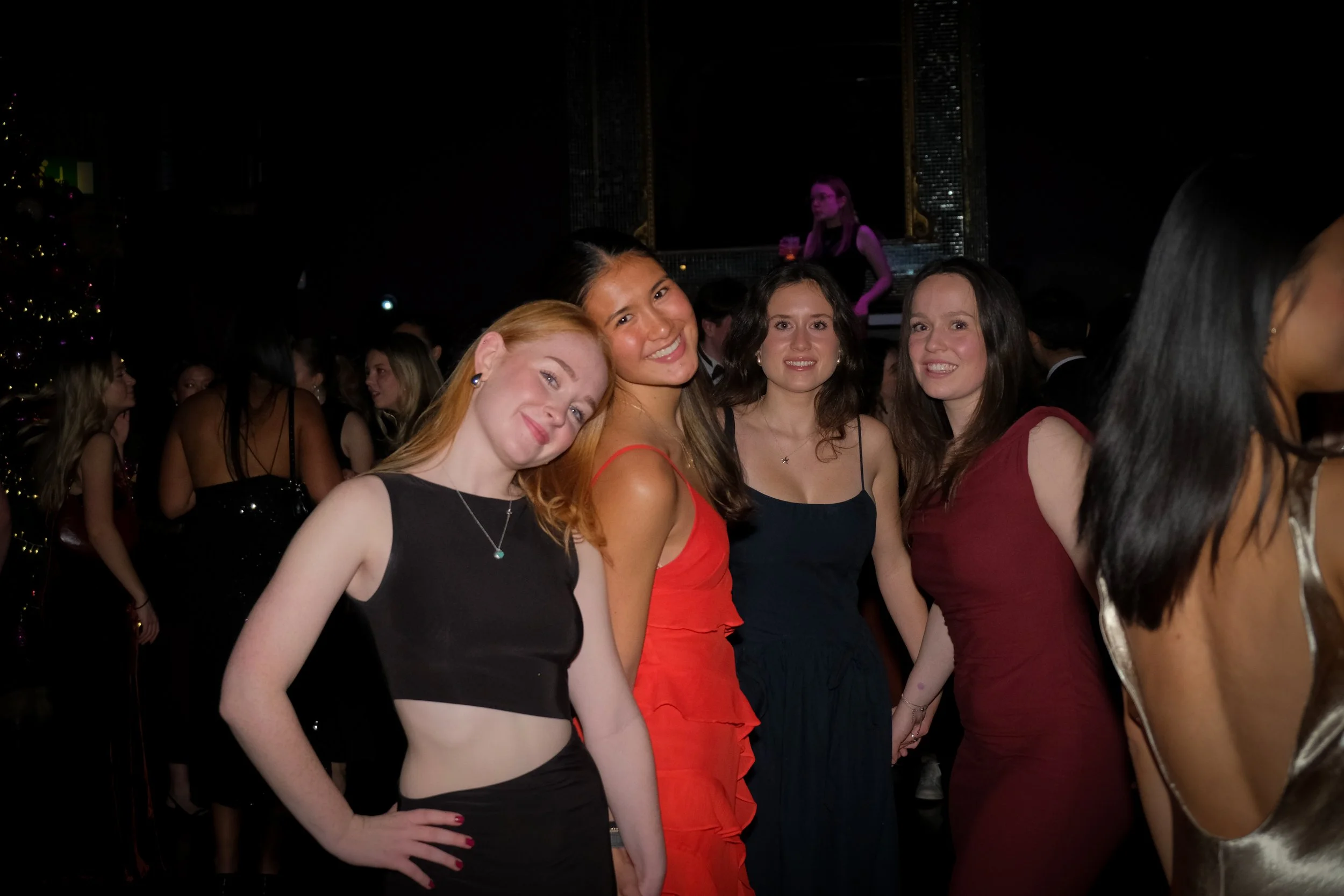 A group of five young women smiling and holding hands at a party or celebration, dressed in elegant evening attire, in a dimly lit venue with other guests in the background.