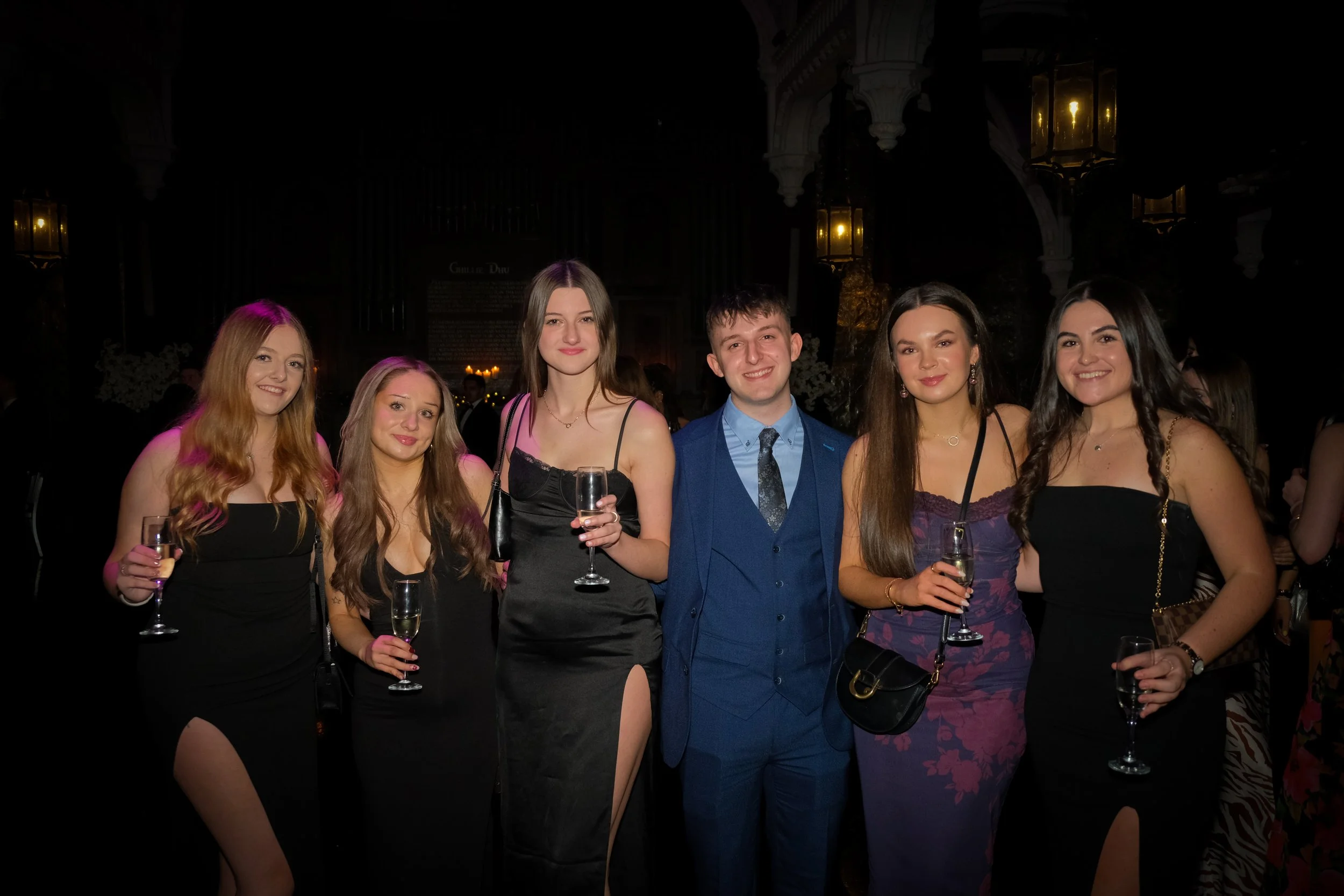 Group of six young adults dressed in formal attire, holding champagne glasses, standing together at an indoor event with dim lighting and elegant decorations.