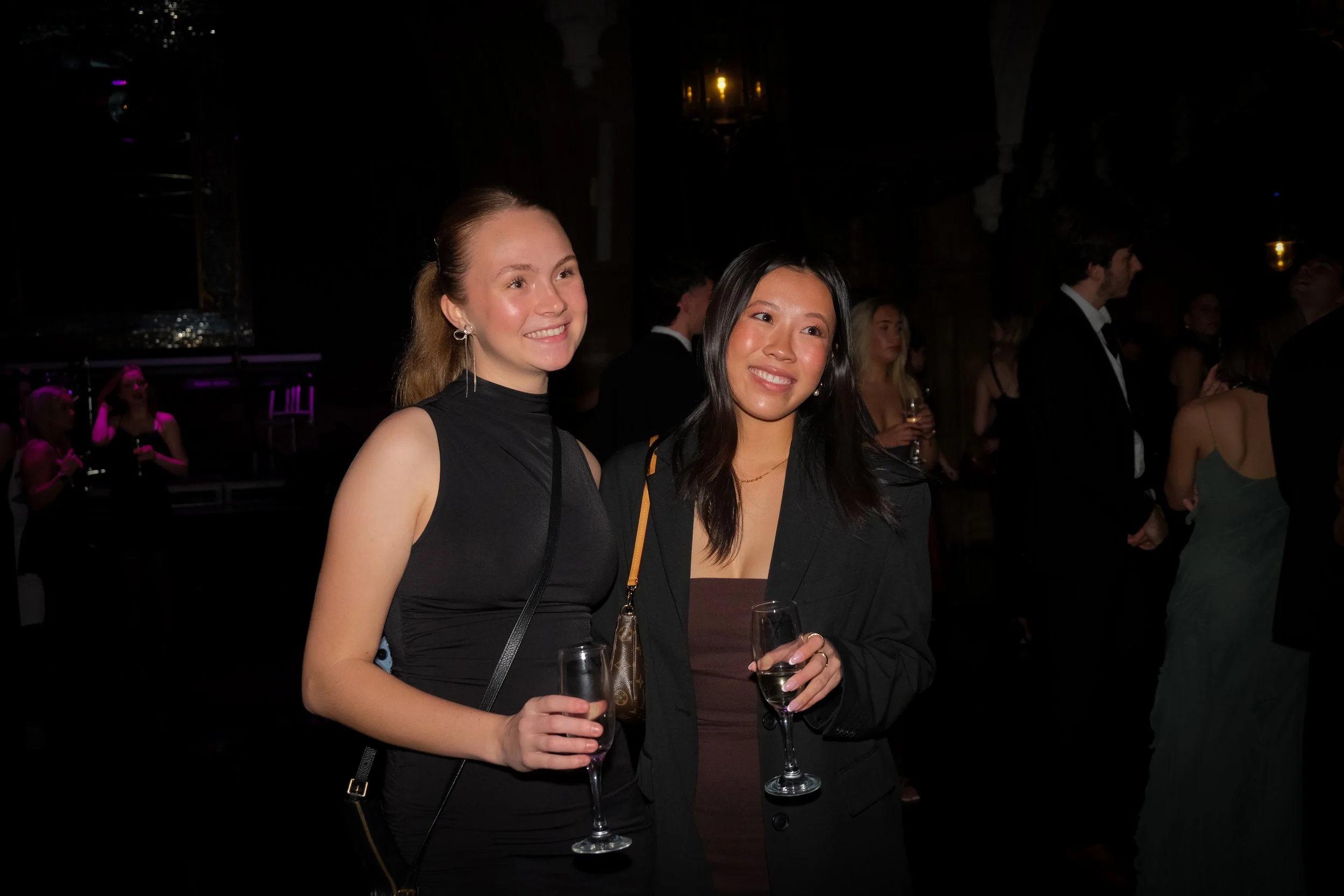 Two women in black outfits holding glasses of champagne at a social event with other guests in the background.
