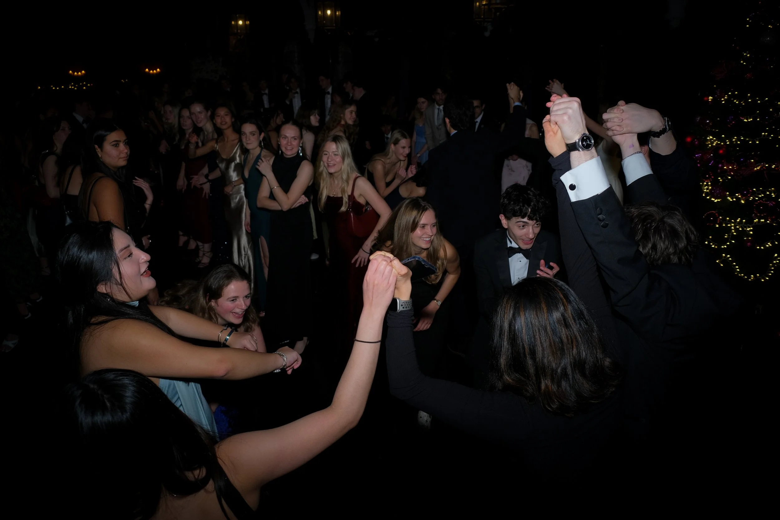 Group of young people dancing and socializing at a formal event or holiday party with Christmas decorations in the background.