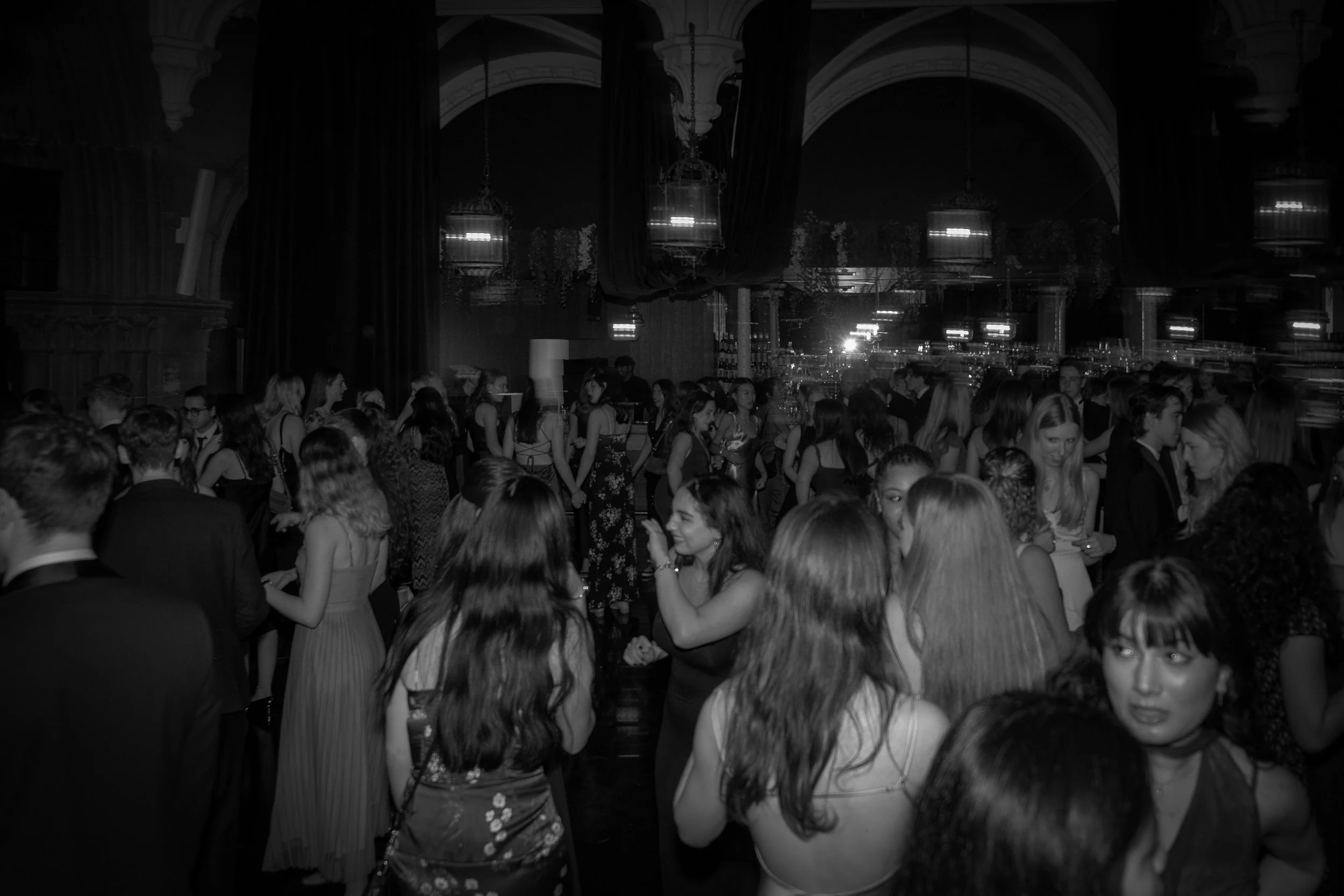 A black and white photo of a crowded indoor party or dance event with many young adults socializing, talking, and dancing in a large, elegant room with arched ceilings and hanging lights.